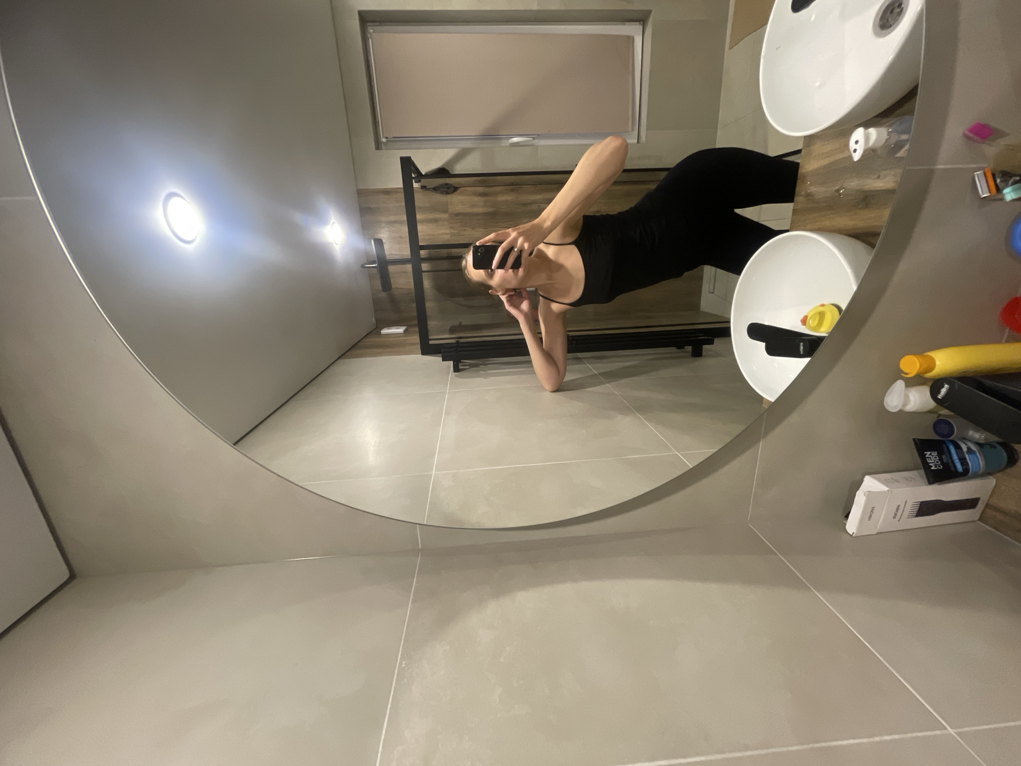This media file depicts a person taking a mirror selfie in a bathroom. The subject, a female, is wearing a black tank top and leggings. She is leaning against a vanity with two sinks. A window with a roller blind is visible in the background. Various toiletries are scattered on the counter and shelf, including bottles, tubes, and a razor. Text visible on one of the tubes reads "MEN CODE". The lighting suggests it is daytime, or the bathroom lights are on. The scene is a modern bathroom with tiled walls and floor. The reflection shows a wide mirror that captures the upper body of the subject and a portion of the bathroom. The overall mood is casual. The location is identified as Подшивалово, Russia.