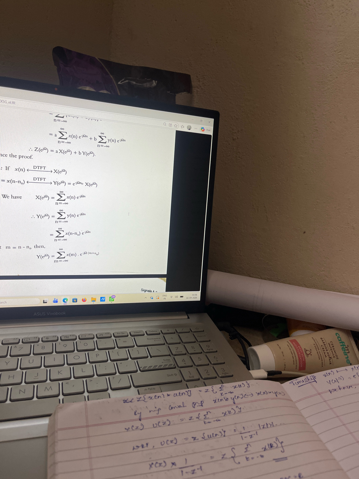 The image shows a close-up view of a laptop screen displaying mathematical equations, possibly related to digital signal processing. On the right side of the laptop, a tube of "Caffeine" brand moisturizer is visible. In the foreground, a notebook with handwritten mathematical formulas is open, placed on top of the laptop's keyboard. The background appears to be a wall with some texture. There are no people visible in the image. The scene is likely indoors, possibly a study or office space, given the presence of a laptop and study materials. The specific location of Vamanjoor, India is not discernible from the visual information provided in the image.