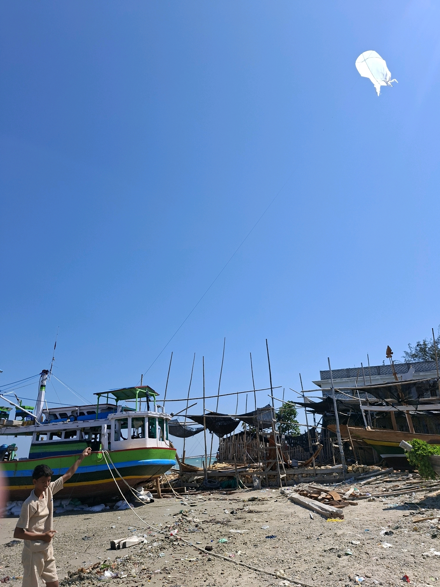 The image depicts a clear blue sky above a beach area in Camplong, Indonesia. In the foreground, a young boy in a light-colored uniform stands on the sand, holding a kite string. He is looking up towards a white kite flying high in the sky. Behind the boy, a large, colorful fishing boat with green, blue, and yellow accents is docked on the shore. The boat is surrounded by scaffolding and construction materials, suggesting it is either under repair or being built. In the background, other boats and structures are visible, along with dense vegetation. The beach itself is covered with sand, debris, and various construction remnants, indicating an active boatyard or repair facility. The overall scene is bathed in bright daylight, casting distinct shadows.
