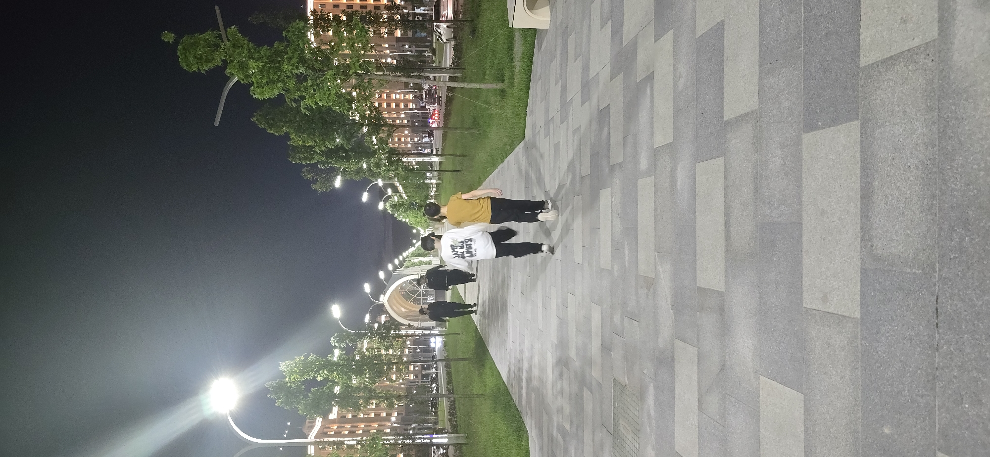 A nighttime scene shows a pedestrian walkway in Grozny, Russia. The walkway is paved with grey rectangular stones and runs alongside a grassy area with trees and street lamps. Buildings are visible in the background. Four individuals are walking along the walkway. One person is wearing a yellow shirt and another a white shirt with writing on it. The area is illuminated by street lamps.