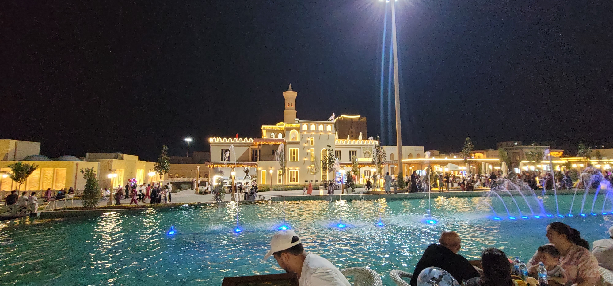 The image depicts a nighttime scene at the newly renovated historical site in the city of Shakpak, Uzbekistan. In the foreground, a wide pool with illuminated blue water jets creates a dynamic fountain display. Several people are seated at tables in the foreground, appearing to be dining or socializing at an outdoor cafe. Their backs are mostly to the camera, obscuring clear details of their faces, though some are clearly visible. A person in a white cap is in the lower-left of the frame, looking down.
The background features a large, elaborately decorated building with an architectural style reminiscent of traditional Central Asian structures, complete with minarets and ornate facades. This building is illuminated by warm, yellow lights and strings of smaller lights that outline its features. Numerous people are walking around in front of the building and along the edges of the pool, suggesting a lively public gathering. The sky is dark, indicating nighttime. The overall impression is one of a well-lit, festive public space designed for entertainment and tourism.