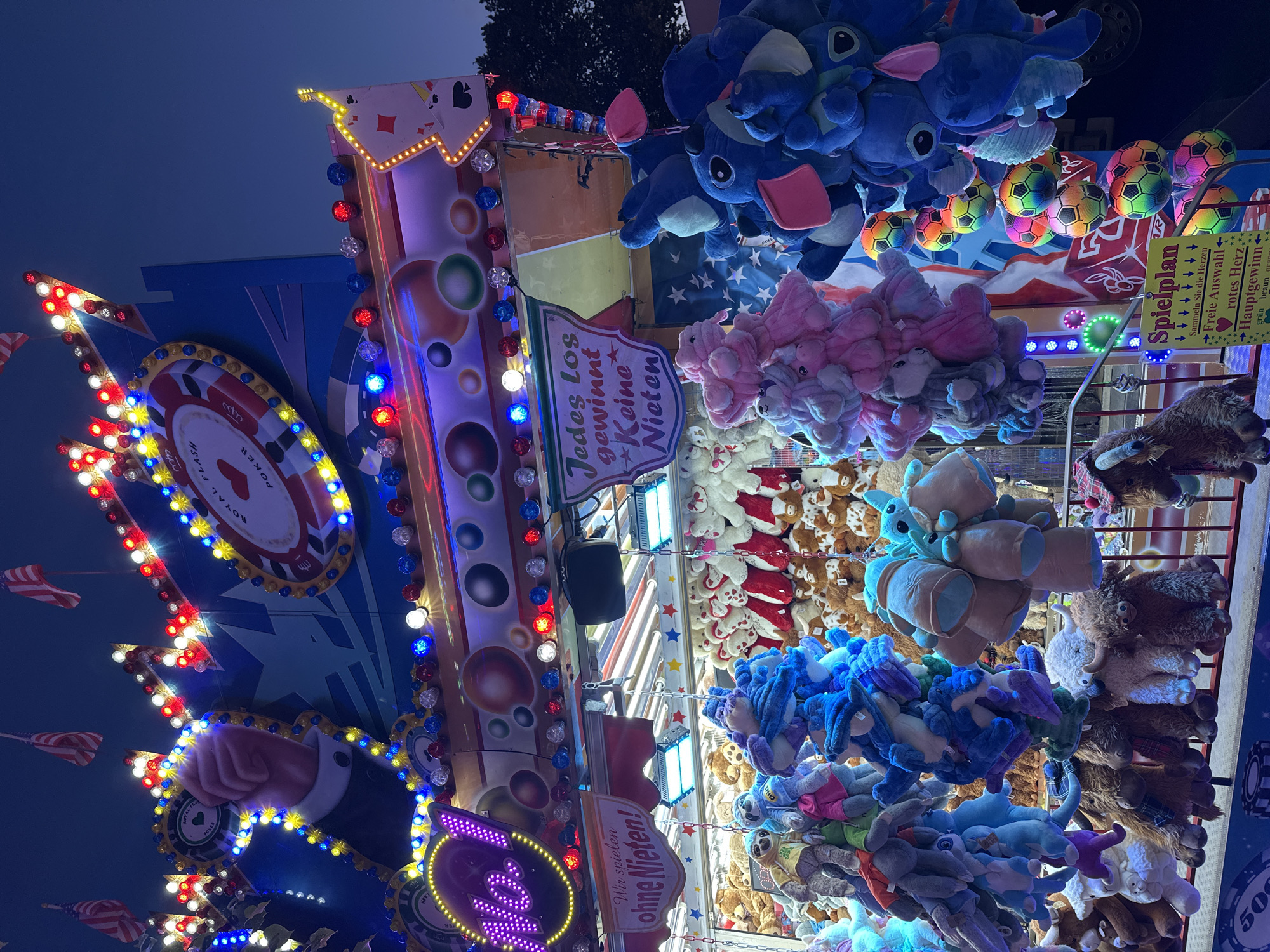 This image captures a brightly lit stall at an amusement fair in Bruchhausen-Vilsen, Germany, after dusk. The stall features a carnival game with plush toys as prizes. The game's facade is adorned with colorful lights and graphics reminiscent of playing cards and poker, including a stylized roulette wheel displaying "ROYAL FLUSH POKER". Large, illuminated signs read "No!" in purple neon and "Wir spielen ohne Nieten!" and "Jedes Los gewinnt - Keine Nieten!" in German, indicating that every ticket wins and there are no losers. Hanging from the stall are numerous plush toys, predominantly in blue, pink, and white, including characters that resemble Stitch from Disney's Lilo & Stitch. Smaller prizes, like colorful soccer balls, are also visible. In the background, more plush toys are displayed on shelves, and American flags are flown from the top of the structure. The overall scene is vibrant and festive, characteristic of a funfair atmosphere.