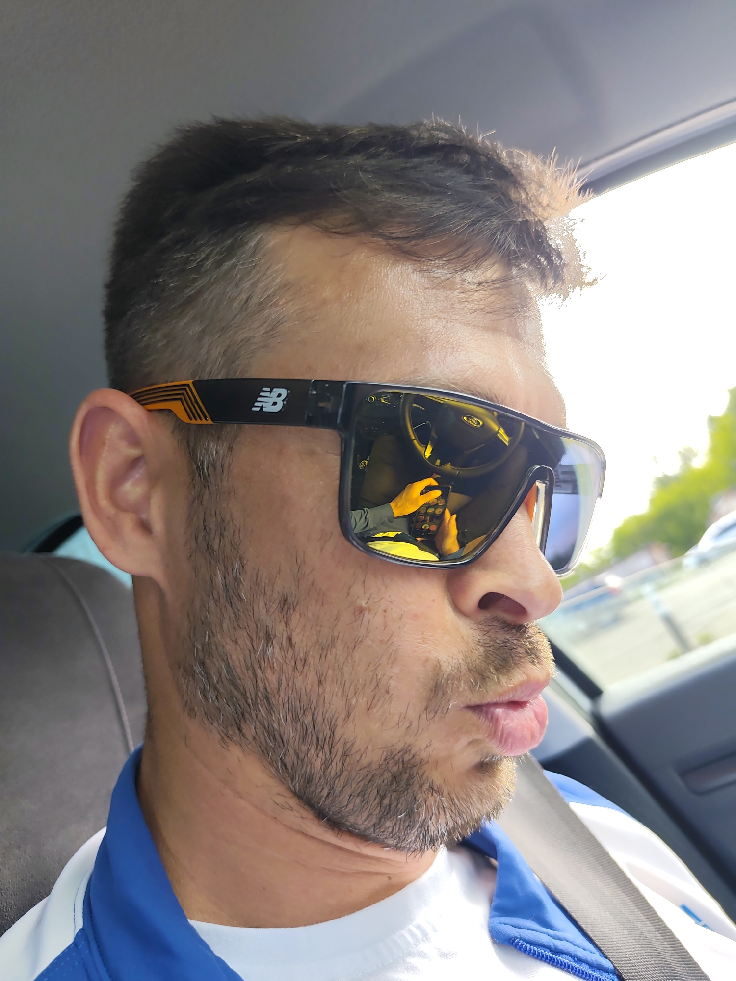 A man with short brown hair and a stubbly beard is sitting in a car. He is wearing a white t-shirt, a blue and white jacket, and a seatbelt. He is also wearing black New Balance sunglasses with orange accents, which reflect the interior of the car, including the steering wheel and his hands holding a cell phone. The background shows a window and what appears to be a parking lot with other cars. The image was taken in the city of Chaikovsky, Russia.