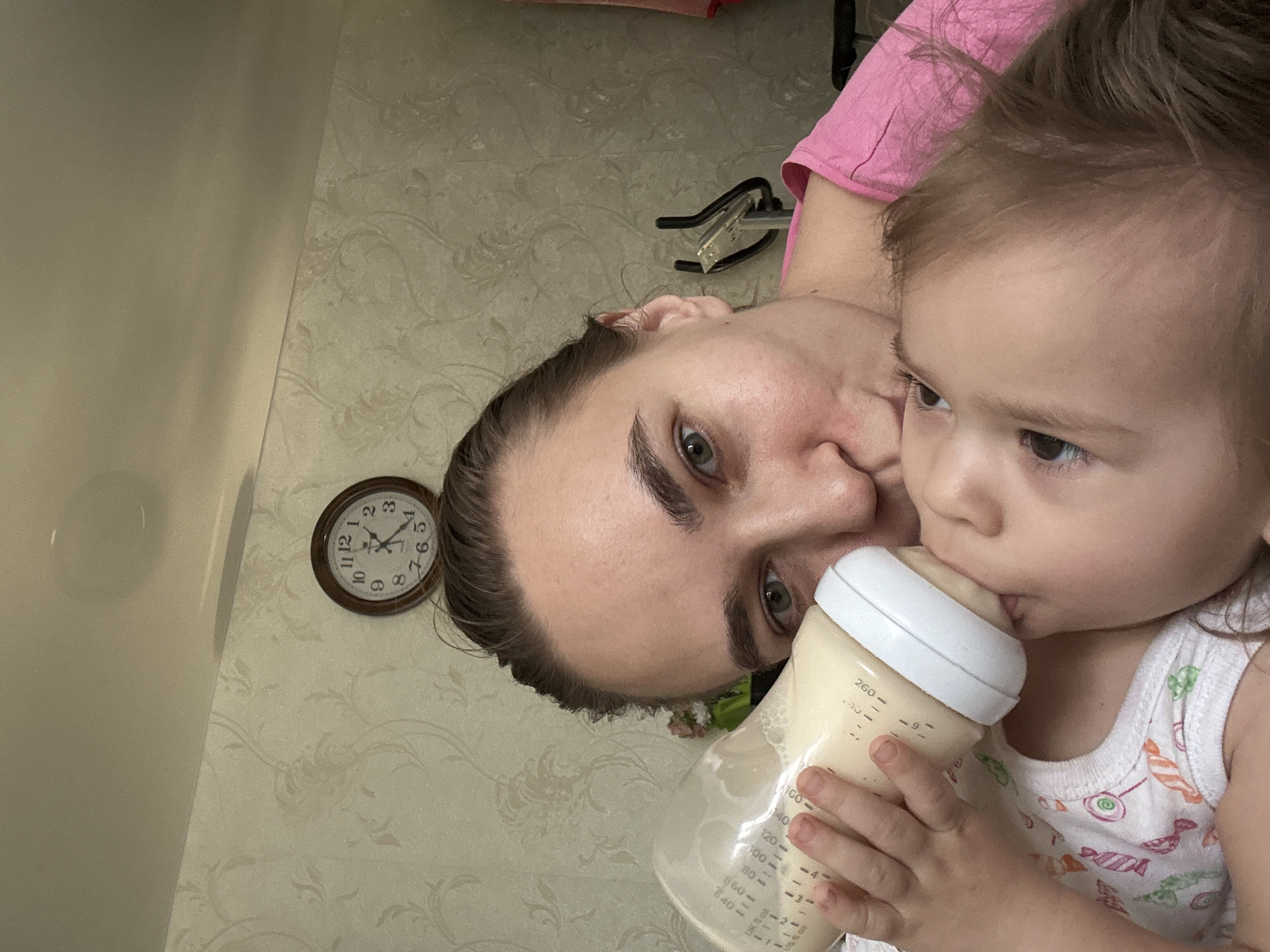 An adult and a young child are captured in a close-up, informal moment indoors in Uva, Russia. The image is taken at an angle, possibly a selfie. The adult, wearing a pink top, has brown hair pulled back, visible green eyes, and looks towards the camera. They are holding a baby bottle filled with a light-colored liquid (likely formula) for the child. The child, with light brown hair, is wearing a white tank top with colorful patterned designs (appearing to be candies or abstract shapes) and is actively drinking from the bottle, their attention focused on it. The adult's face is close to the child's, showing an intimate and nurturing interaction.
In the background, a patterned wallpaper with a subtle floral or swirl design is visible on one wall, while an adjacent wall or surface is plain and light-colored. A round wooden-framed clock on the patterned wall shows the time as approximately 1:50. Another object, possibly a stapler or a specific tool, is partially visible on the patterned wall behind the adult's shoulder. The baby bottle has clear measurement markings: "260," "160," "140," "120," "100," "80," "60," "40" in milliliters, and corresponding ounce markings from "9" down to "1." Faintly visible text includes "ml," "oz," "UK," and part of "Litres." The overall atmosphere is calm and focused on the feeding activity.