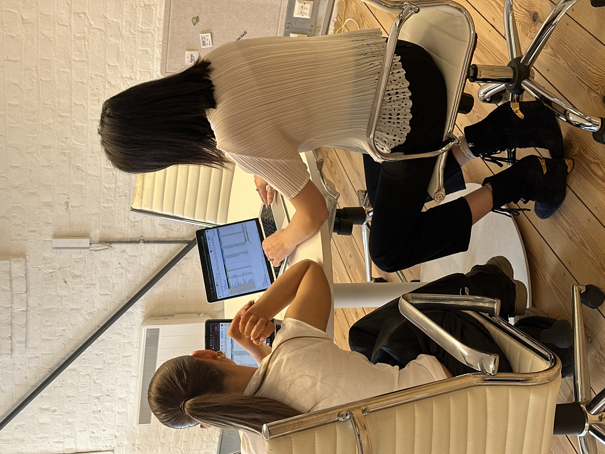 Two individuals are seated at a white round table in an indoor office space located in the city of London, United Kingdom. The background consists of a white-painted brick wall featuring a diagonal dark metal structural beam and a white wall-mounted rectangular radiator. A vertical white conduit pipe and electrical outlet are visible on the right wall. The floor is made of light-toned wooden planks.
The individual on the left, viewed from behind, has light brown hair styled in a high ponytail. They wear a white short-sleeved top, dark trousers, and gold hoop earrings. Their hands are clasped on the tabletop near a dark-framed tablet device displaying a user interface. This individual is seated in a cream-colored office chair with chrome armrests and a wheeled base.
The individual on the right, also viewed from behind, has straight, dark, shoulder-length hair. They wear a light-colored, possibly off-white, pleated short-sleeved top with a distinct scalloped and perforated lace-like hemline, paired with black trousers. Their hands are positioned over the keyboard of a silver laptop, which is open on the table and displaying a spreadsheet application with multiple rows and columns of data. This individual is seated in a similar cream-colored office chair with chrome elements. Their footwear consists of dark blue suede lace-up ankle boots with gold-colored heels and heel accents. Both individuals are actively engaged with their respective electronic devices.