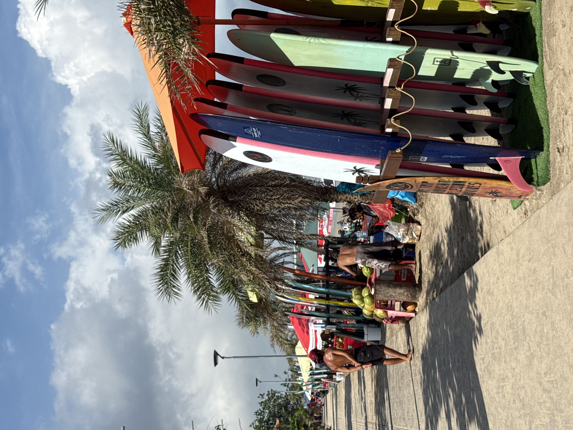 The image shows a street scene in Kuta, Indonesia. A rack displays several surfboards of various colors and designs under an orange umbrella and a palm tree. A shirtless man in black shorts stands on a concrete path next to the rack, his back to the camera. Further down the path, other people are gathered around what appears to be a fruit vendor's cart with coconuts. The sky is partly cloudy. The general atmosphere is of a tropical beach town.
