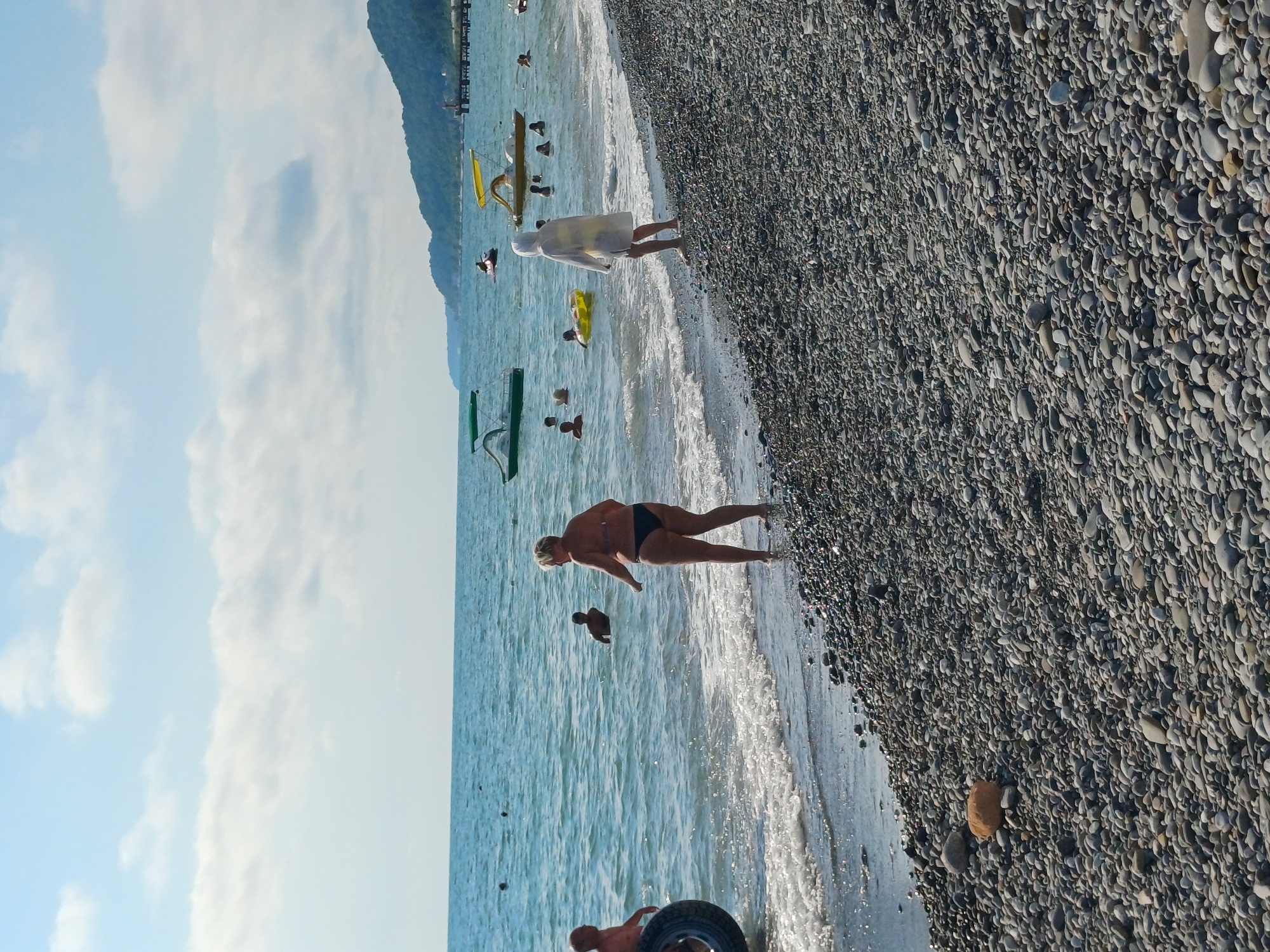 This is a media file of a beach scene in Sochi, Russia. The image is oriented vertically. The foreground is a pebble beach with a person in a black bikini standing near the water's edge. Behind them, several other people are in the shallow, turquoise water, some with inflatable toys. In the background, a person in a white hooded outfit walks towards the water, with a yellow inflatable structure nearby. A pier or jetty extends into the sea in the far distance, with a dark green object and a yellow flag on it. The sea is calm, with gentle waves lapping the shore. The sky is bright blue with scattered white clouds. The overall mood appears to be a sunny, pleasant day at the beach. There is no visible text in the image.
