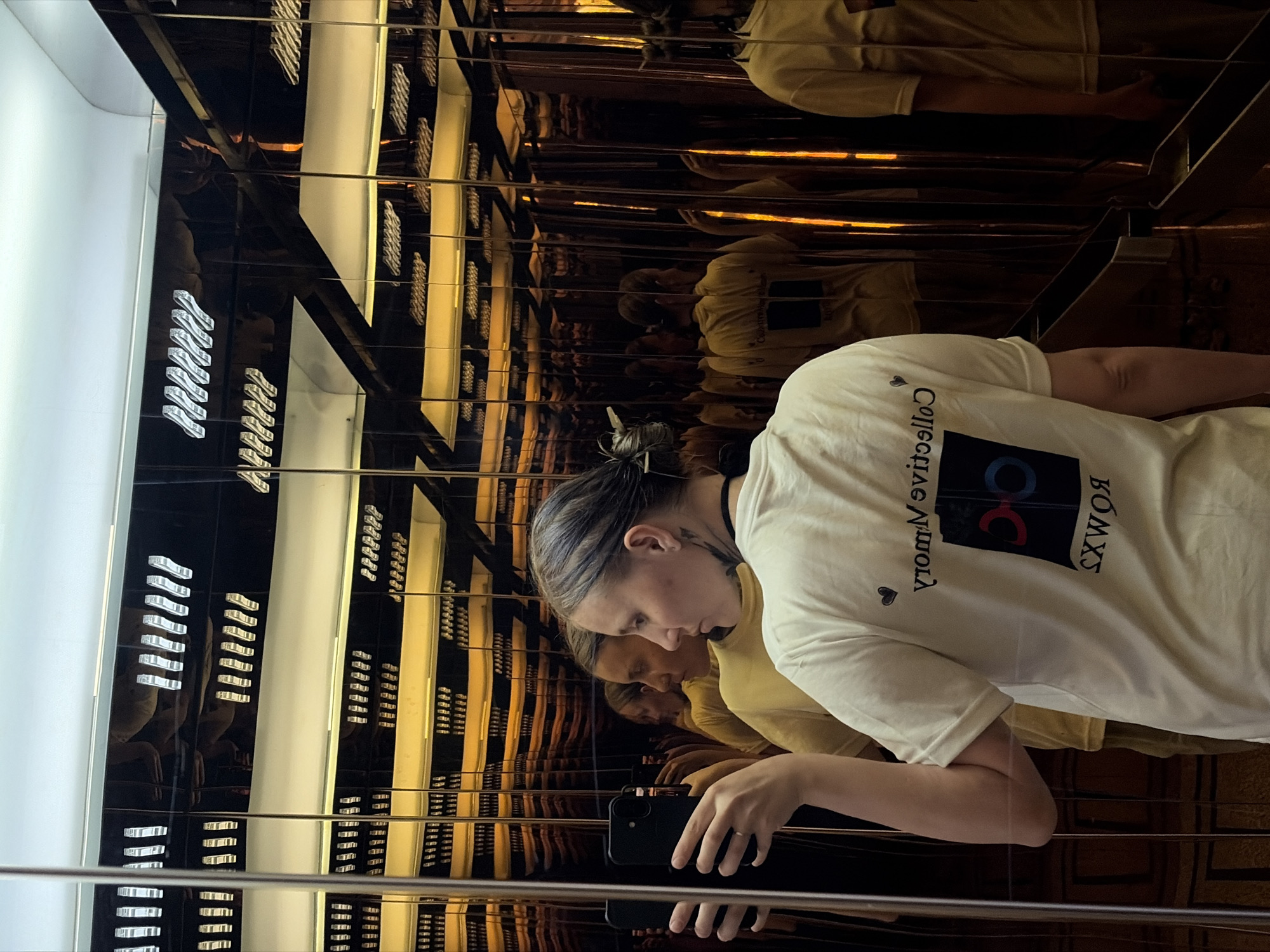 A person is depicted in an indoor setting, appearing to take a photograph of their reflection with a black smartphone. The image is oriented vertically, showing the subject from the chest up, with their head slightly bowed as they look at the phone screen. They have light-colored hair pulled back and a visible tattoo on their neck. The person is wearing a white t-shirt with text and a logo printed on the back. Due to the mirrored surface, the text reads backwards as "vroM eviteclloC" (Collective Memory) and "SXWOQ", alongside a square and circle graphic, and a small heart icon. A ring is visible on one of their fingers.
The environment is highly reflective, featuring numerous golden-hued, possibly mirrored or polished metal, vertical surfaces that create an illusion of infinite depth and repetition. Bright, linear strip lights are reflected countless times, creating a striking visual pattern across the scene. Other reflections show a blurred silhouette of another person in the background and multiple instances of the main subject and their phone. The overall ambiance is modern and visually complex due to the extensive use of reflective materials and artificial lighting. This unique setting is located in Đà Nẵng, Vietnam, possibly within an art installation, a hotel lobby, or a contemporary building.