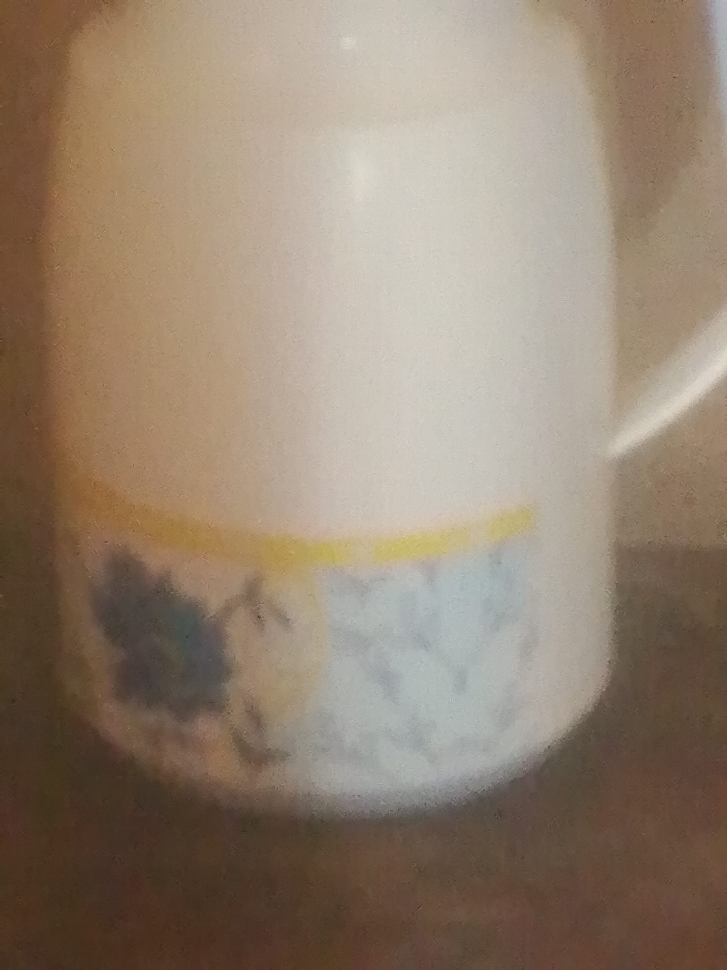 The image shows a close-up of a white thermos flask with a decorative band around its base. The band features a pattern of light blue and grey abstract floral or leaf shapes, with a thin yellow line separating it from the white body of the flask. The background is out of focus and appears to be a dark, textured surface, possibly a wooden table. The lighting is soft and diffused, suggesting an indoor setting. There is no text visible in the image. The image does not depict any people or specific activities, focusing solely on the object. The setting appears to be domestic or a simple interior space, with no clear indicators of time of day or weather. The overall impression is that of a simple product shot.