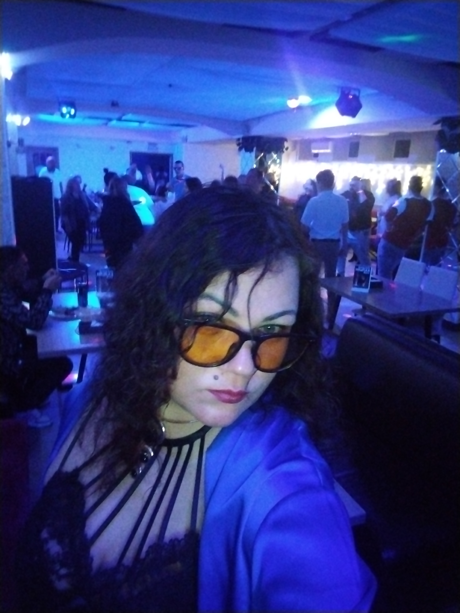 This media file captures an indoor scene at what appears to be a bar, club, or restaurant in Chaikovsky, Russia, during the evening or night. The room is dimly lit, predominantly by blue artificial lighting, with some stage or disco lights visible on the low ceiling, casting a cool tone over the entire space.
In the foreground, a woman with long, dark, curly hair is taking a selfie. She is wearing large, orange-tinted sunglasses, a black lace top with prominent strap details, and a blue jacket. Her lips are painted a deep red, and a small mole or piercing is visible above her upper lip. She holds the camera low, looking upwards slightly.
The background is crowded with numerous other people, mostly adults, who are socializing. Some are standing, while others are seated at tables or in booths. Drinks in glasses are visible on a table to the left. The general atmosphere suggests a lively social gathering or party. The people in the background are less distinct due to the dim lighting and slight blur, but they appear to be casually dressed. Overhead, several light fixtures, including what looks like a projector or moving head light, are suspended from the ceiling. A small, illegible piece of text on a stand or menu is visible on one of the tables in the mid-ground.