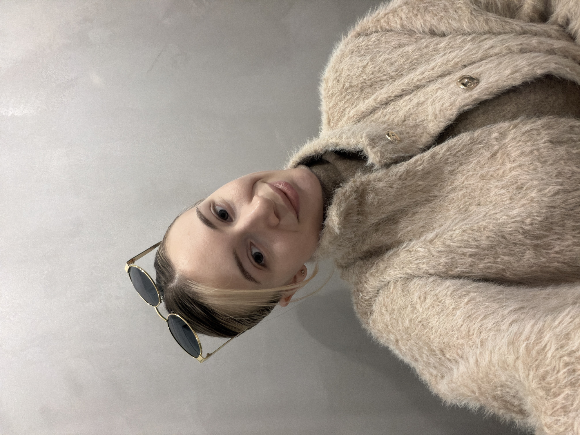 A woman with light skin is wearing a beige, fuzzy coat and sunglasses with gold frames on her head. She is looking directly at the camera and smiling slightly. The background is a plain, gray wall. There is no indication of the location being Plovdiv, Bulgaria in this image.