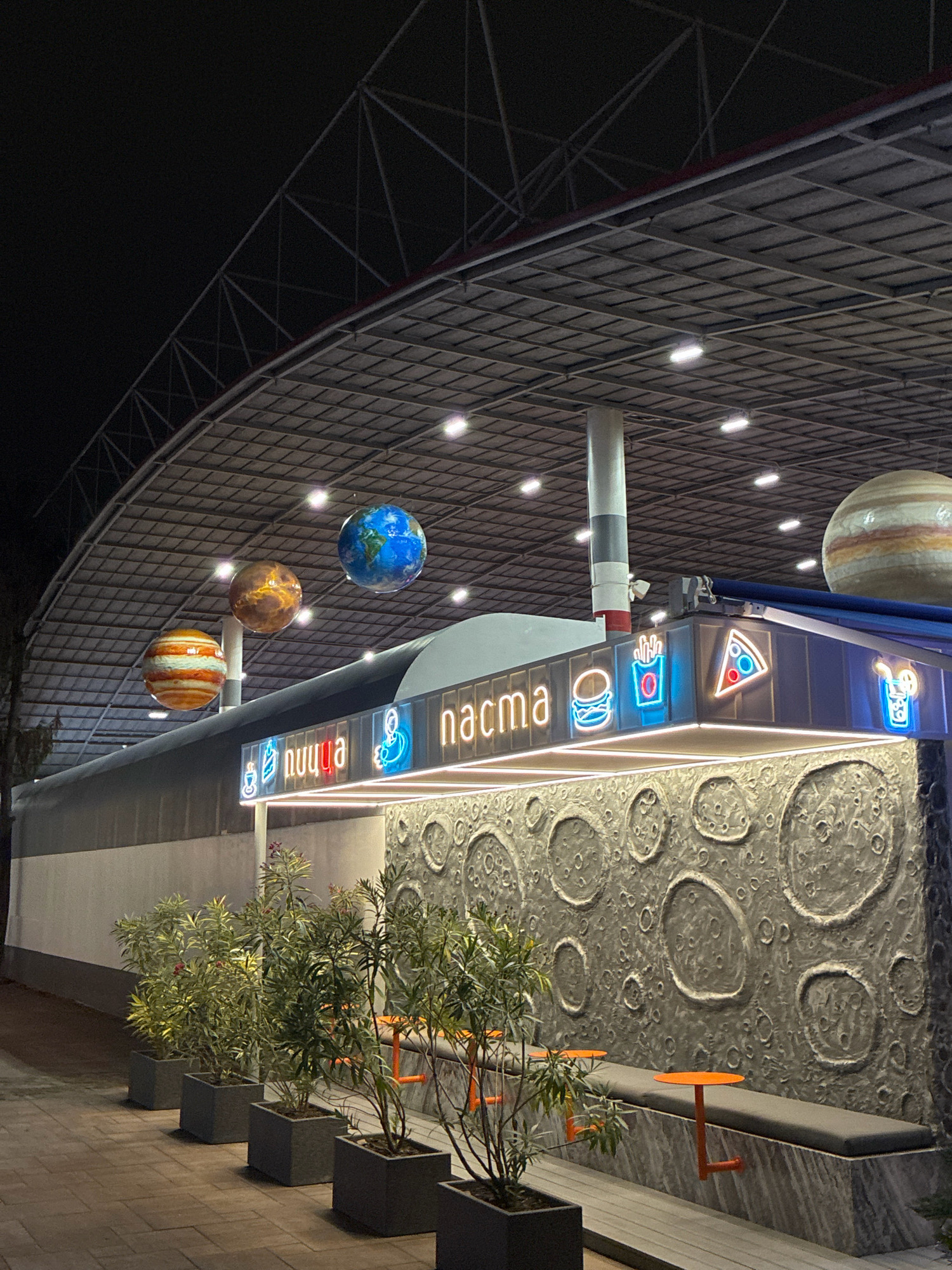 The image captures a night-time view of a unique outdoor dining area designed with a space theme. The scene is located in Sochi, Russia, likely a part of an entertainment venue or a themed cafe.
The foreground shows several evenly spaced, dark grey square planters, each containing a small, leafy green plant with some red blossoms. These planters are arranged on a paved walkway made of reddish-brown cobblestones.
Behind the planters is a long, built-in seating area with dark grey cushions. Orange, L-shaped tables are attached to the wall at intervals along the bench. The wall behind the seating is a prominent feature, decorated with a textured grey relief resembling the surface of the moon, complete with various sized craters.
Above the seating area is a modern food stand or counter. It has a dark blue facade adorned with illuminated neon signs. To the left, a neon sign displays the word "nuuqa" in pink and blue, with a stylized figure to its right. Further to the right, neon signs depict a hamburger, french fries in a red box, a slice of pizza with red and blue toppings, and a blue drink with a straw and lemon slice. The word "nacma" is visible in neon lights on the facade.
Suspended from the curved, grid-like ceiling structure above the stand are several large, spherical decorations resembling planets. From left to right, these appear to be Jupiter (orange and white stripes), a planet with a brown and orange marbled surface (possibly Mars or Venus), and Earth, clearly depicting continents and oceans. A large, striped planet, possibly another Jupiter, is partially visible in the upper right corner of the image.
The ceiling itself is a dark, angled structure with a metal grid pattern, illuminated by scattered warm white lights. The overall ambiance is futuristic and playful, with the space theme extending from the decorations to the textured wall and neon signage. The lighting creates a dramatic and inviting atmosphere in the dark. No people are visible in the image.
