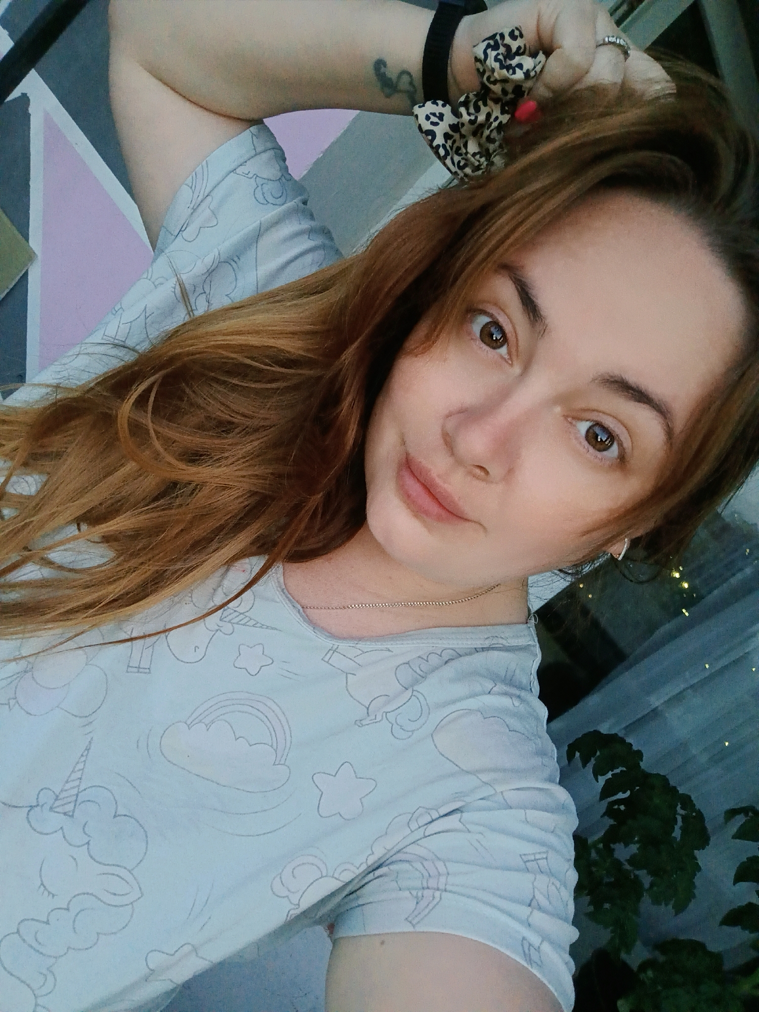 A young woman with long, auburn hair and brown eyes is shown in a close-up, selfie-style shot. She is wearing a light blue t-shirt with a pattern of unicorns, clouds, and stars. Her left arm is raised, and she is holding a leopard-print scrunchie with a red bead in her hand. She has a tattoo on her left forearm and is wearing a ring on her right ring finger. The background appears to be outdoors, with a wall featuring a geometric pattern of pink, gray, and white sections. In the lower right corner, some green foliage is visible, and there are small, blurred lights in the distance, suggesting it might be evening. The image was taken in the city of Chaikovsky, Russia.