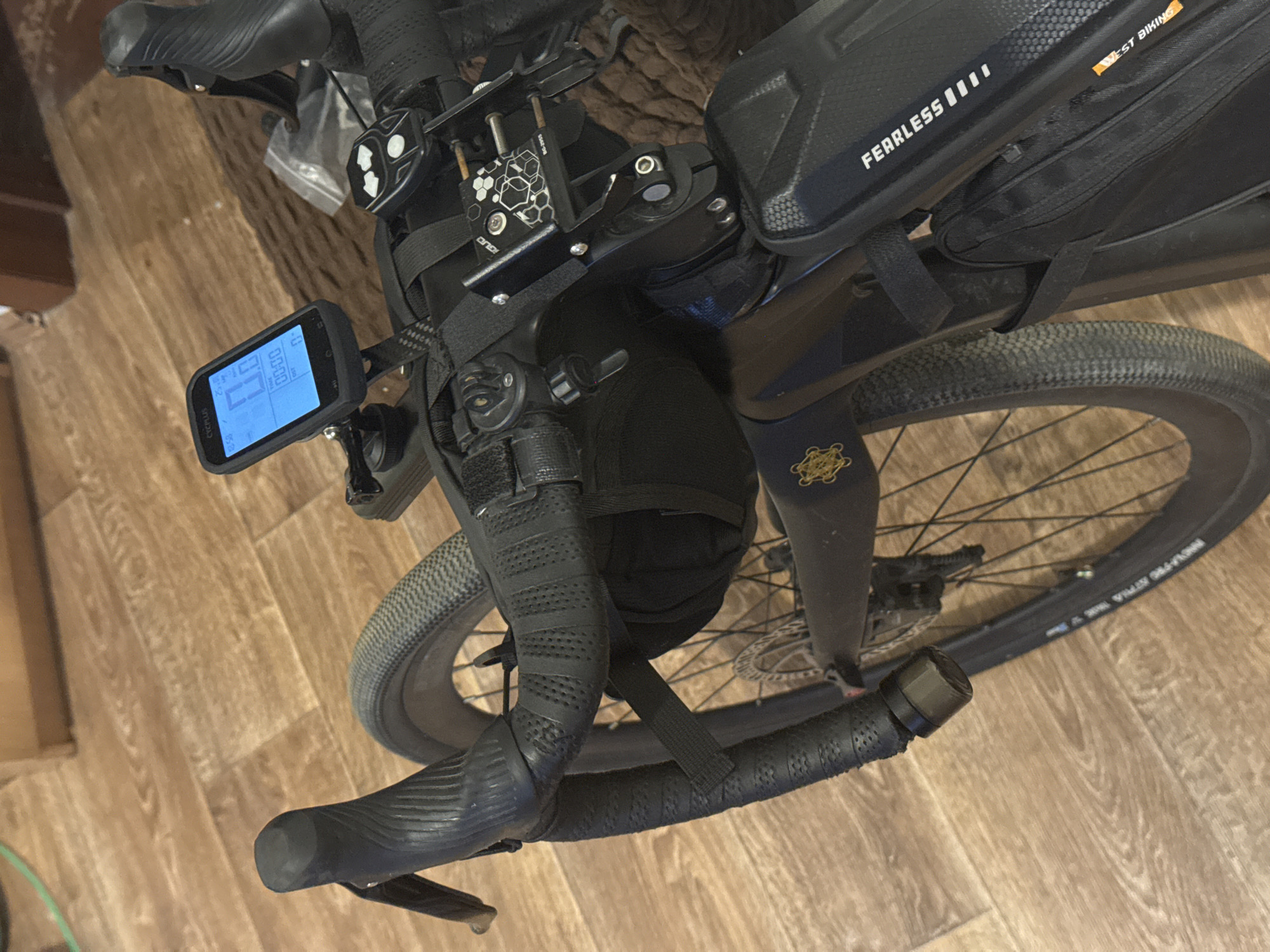 A close-up shot of the front of a bicycle in Ново-Никольское, Russia. The bicycle has a black frame and is equipped with accessories for long-distance riding, including a handlebar bag and a top tube bag labeled "FEARLESS". A GPS device is mounted on the handlebars. The tires are wide and knobby, suitable for off-road riding. The handlebars are wrapped in black tape. The image suggests a bicycle prepared for a bikepacking or touring adventure.