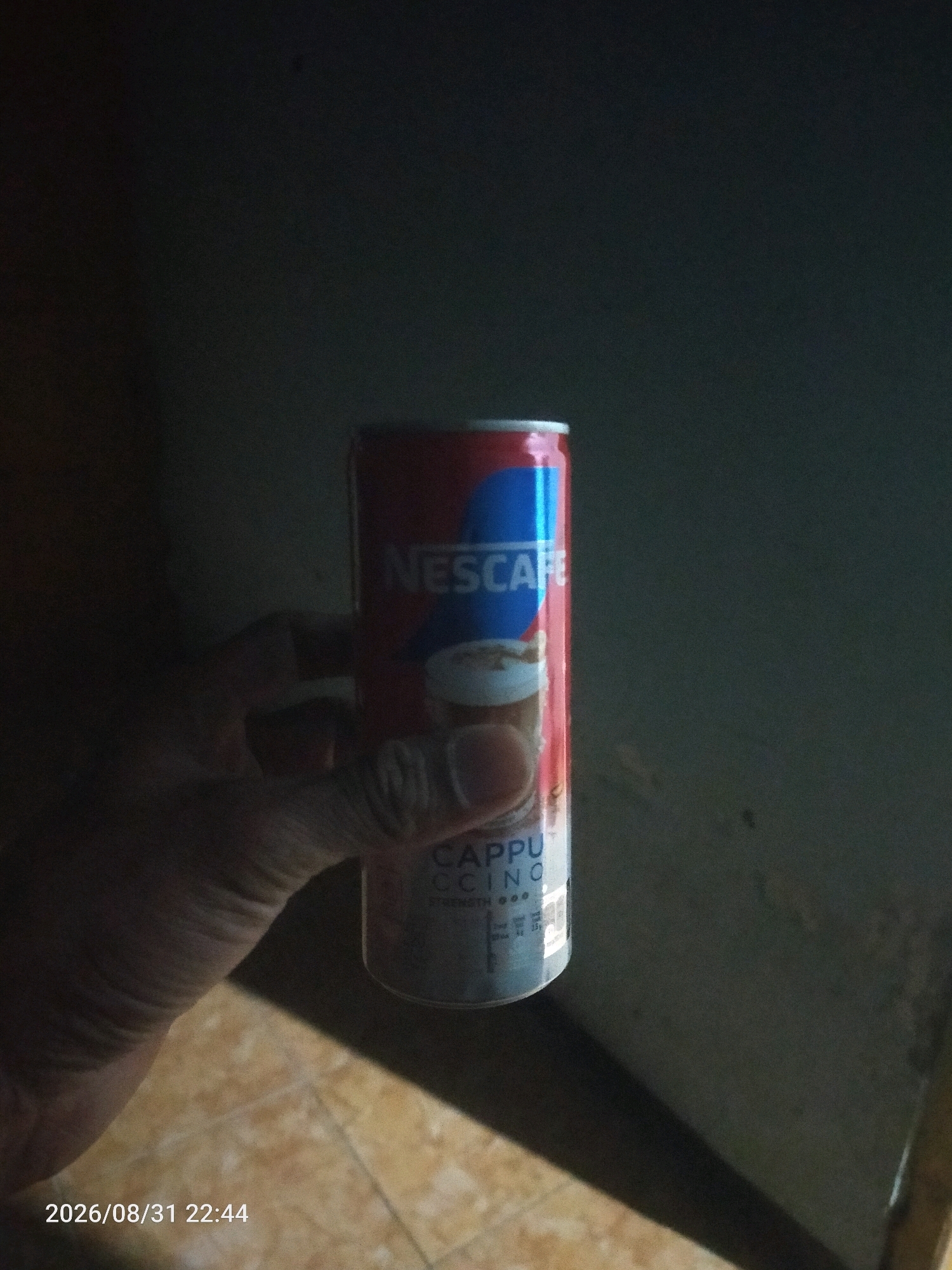 An individual's left hand is holding a red and blue Nescafe Cappuccino can. The can is cylindrical and has a white rim at the top and bottom. The Nescafe logo is prominent on the can. The background is dark and out of focus, with a hint of a tiled floor in the lower left quadrant. A timestamp "2026/08/31 22:44" is visible in the lower left corner. The image appears to be taken indoors, with artificial lighting. The location is Jati Wates, Indonesia.