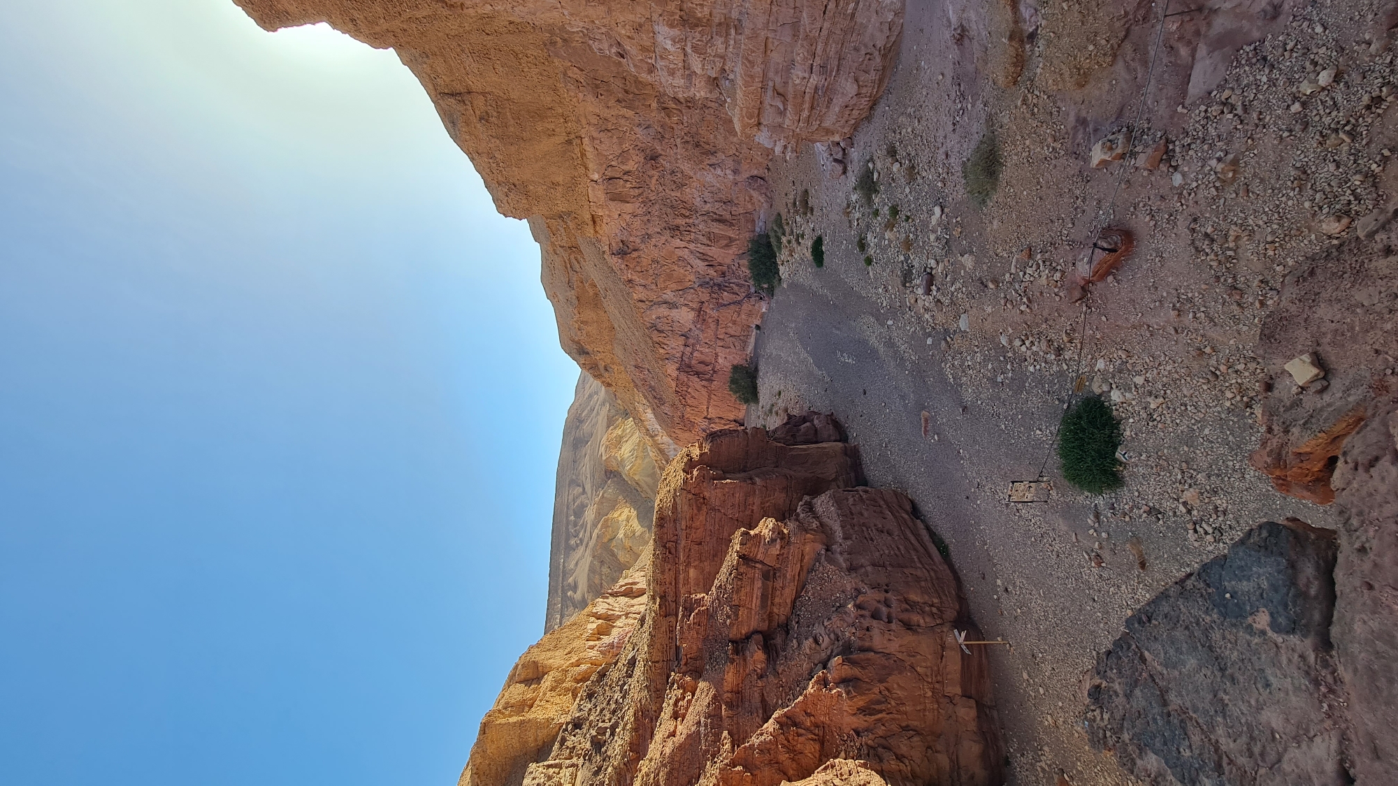 The image depicts a desert canyon, likely in the Hevel Eilot Regional Council, Israel. The canyon walls are composed of reddish-brown rock formations. The canyon floor is a mix of gravel and sand. There are sparse patches of green vegetation. A rope with some metallic objects is stretched across the width of the canyon. A blue sky is visible above one canyon wall.