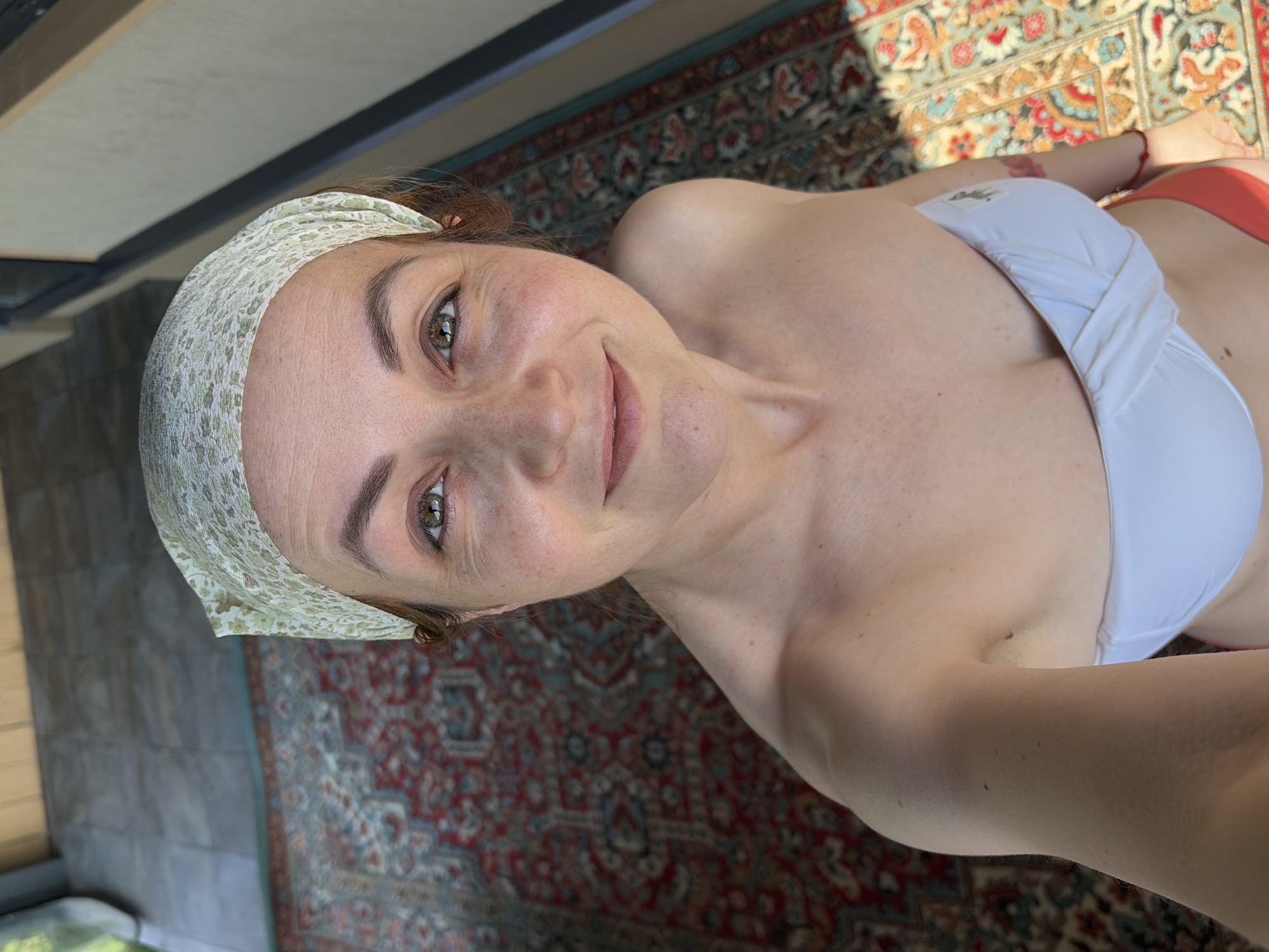 A woman with a white patterned headscarf is positioned in an indoor setting in what appears to be Chaikovsky, Russia. She is wearing a white bikini top and what looks like orange bikini bottoms. She is in a well-lit room with a patterned rug visible. The lighting suggests daytime. The woman is holding the camera at a high angle and is smiling slightly. The background includes a glass door or window with a partially visible sill, next to a patterned rug.