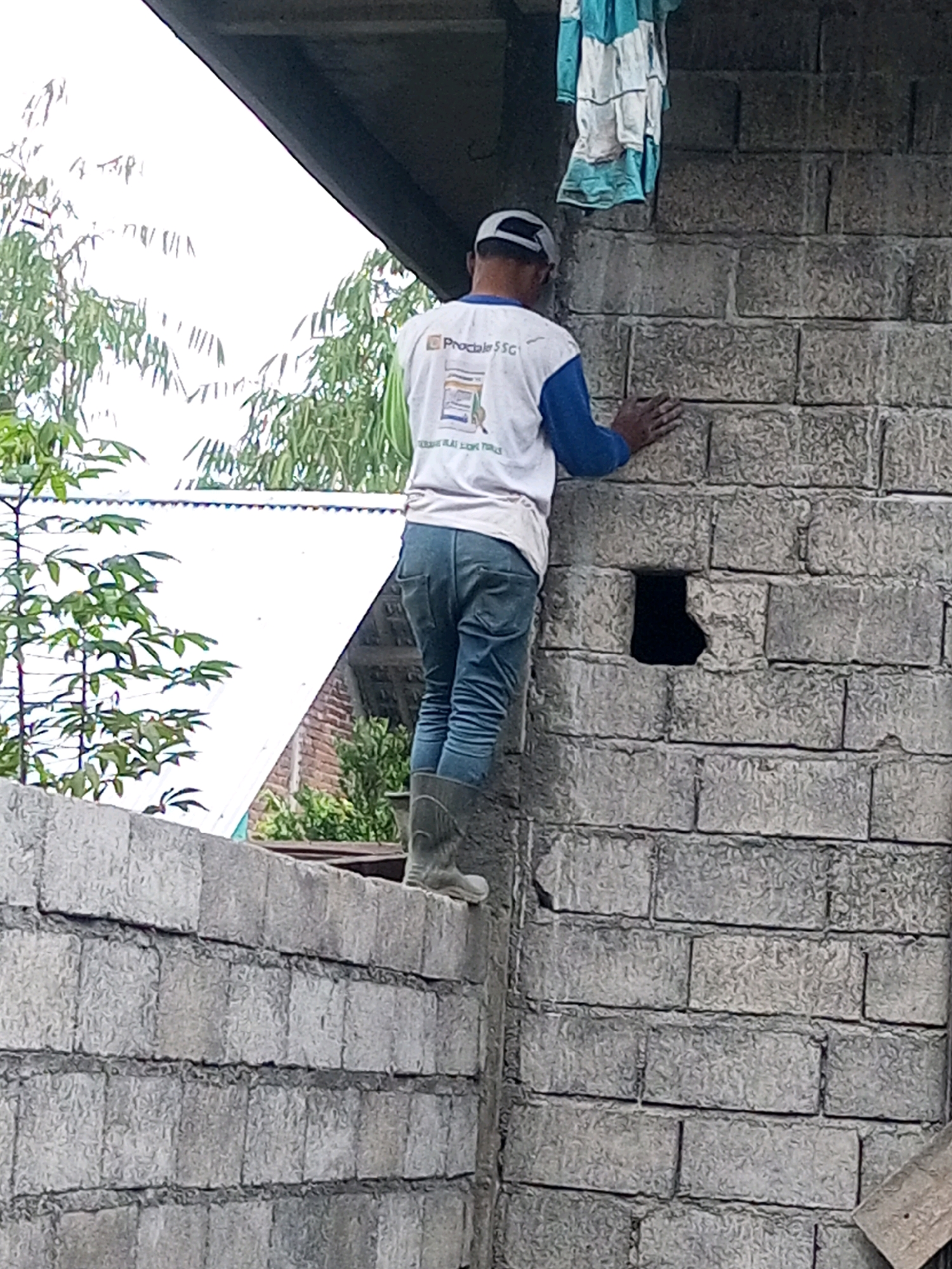 In Andongsili, Indonesia, a person wearing a white t-shirt with blue sleeves and dark jeans stands on a wall, placing their right hand on a cinder block wall. They are also wearing grey rubber boots. A blue and white striped shirt hangs on the wall behind them. The person is looking down at the wall. The background shows trees and a white roof.