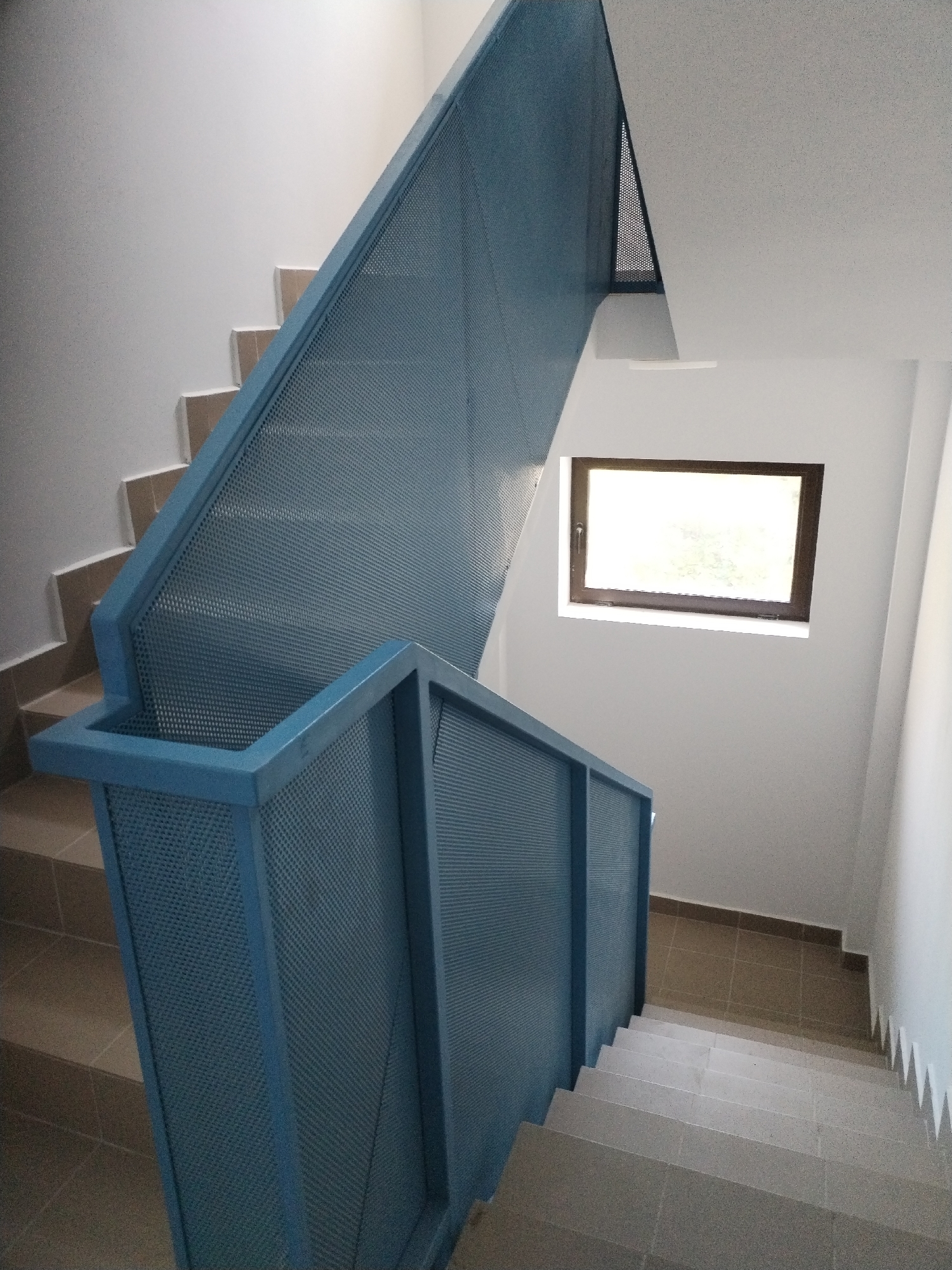 The image shows an interior view of a modern staircase with blue perforated metal railings. The stairs themselves are made of light-colored, possibly concrete or stone, material with a subtle speckled pattern. The walls are painted white. A small, rectangular window with a dark frame is visible on the right wall, admitting natural light. The floor at the bottom of the stairs also appears to be tiled in a neutral, earthy tone. The overall scene is clean and minimalist, suggesting a contemporary architectural design. The lighting is bright, likely from the window and overhead sources. There are no people or explicit indications of a specific location within a city, as the environment appears generic.
