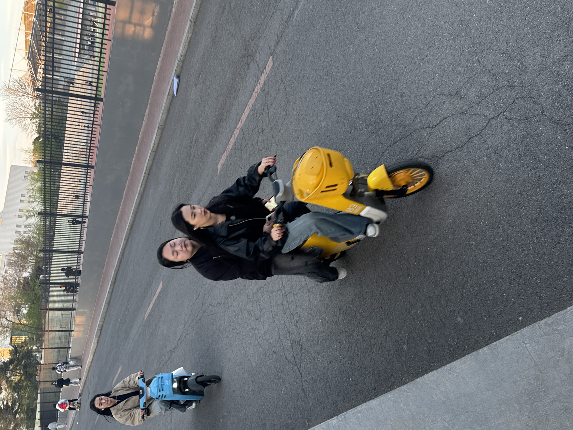 Two young women are riding a yellow electric moped on a paved road. The woman in the front is wearing a black leather jacket and the woman in the back is holding a phone. To the left, a person is riding a blue electric moped. A fence and trees line the left side of the road. In the background, a large building is visible. The location is Zhongtun, China.