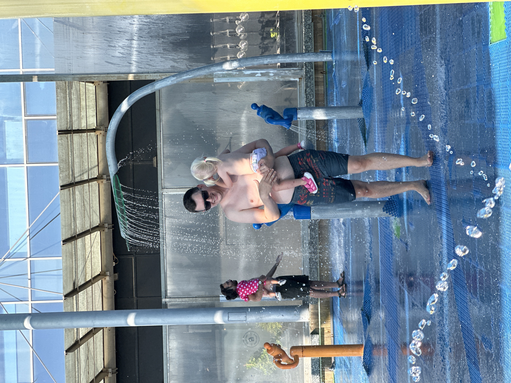 A group of people are enjoying a splash pad on a sunny day. A man in the foreground, shirtless and wearing patterned swim trunks, holds a young girl with blonde pigtails on his hip. Both are in focus and smiling. The girl is wearing a diaper and pink sneakers. Behind them, another man, also shirtless, is holding a toddler wearing a pink shirt with white polka dots and black shorts. The toddler is facing away from the camera and looking towards the man. In the background, there are various water features, including a curved pipe with water spraying from it, and other fixtures that spray water upwards. The splash pad itself is a blue surface with a textured pattern, wet from the water play. The setting appears to be outdoors, with a modern building with large windows and a metal roof structure visible in the background. The overall scene depicts a casual, recreational activity. This image was taken in Hod HaSharon, Israel.