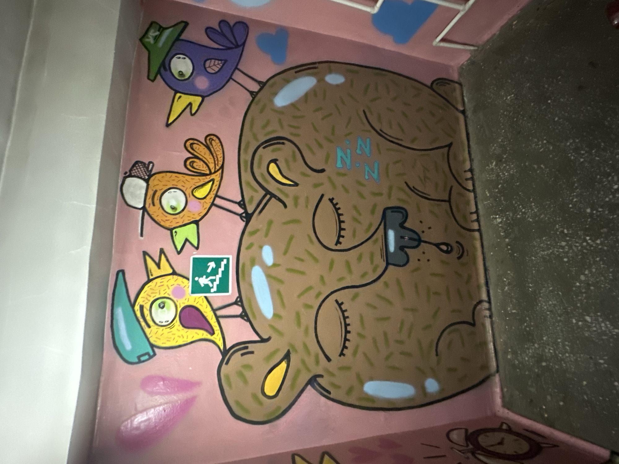 The image shows a colorful graffiti mural on a pink wall, possibly in a public space like a staircase or hallway. The mural features a large, sleeping brown bear with its eyes closed and a drop of drool falling from its mouth. The bear is adorned with numerous small green lines, resembling fur or straw. Perched on the bear's head are three cartoonish birds. The bird on the left is yellow with a teal cap and is holding a green exit sign. The middle bird is orange, wearing a white baseball cap, and appears to be talking to the yellow bird. The bird on the right is purple, wearing a green hat, and is looking to the right. The wall also has pink heart shapes and blue clouds painted on it. To the far left, a red alarm clock with radiating lines is visible. The bottom of the image shows a concrete floor. The overall style of the graffiti is playful and whimsical. This mural is located in Biysk, Russia.