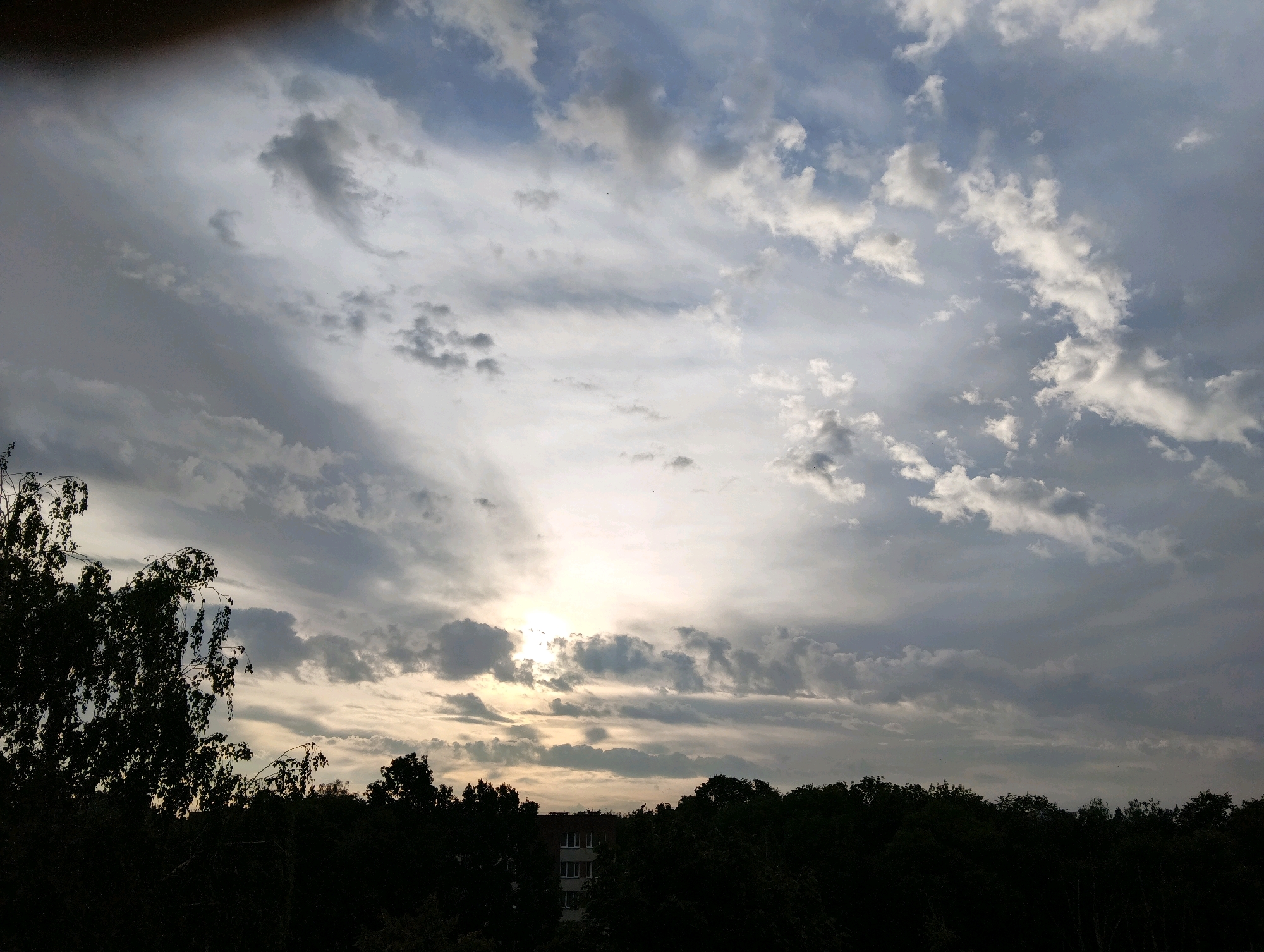The image shows a cloudy sky over a silhouetted treeline with a building visible in the distance, taken from Hrodna, Belarus. The sun is partially obscured by clouds, casting a diffused light across the sky. The clouds are a mix of grey and white, with some appearing darker and more textured than others. The treeline is dense and dark, obscuring any details of what lies beyond. A multi-story building with visible windows can be seen peeking through the trees in the center-right of the frame. The overall impression is one of a slightly overcast day in an urban or suburban setting.