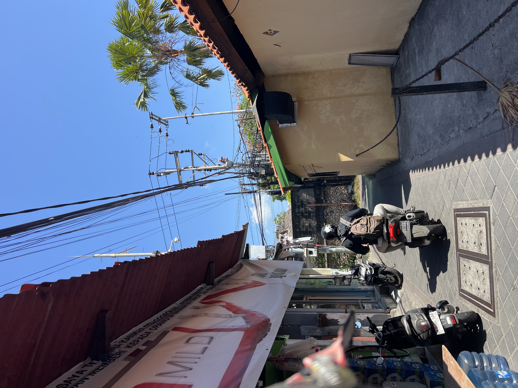 In Kuta, Indonesia, a person wearing a black jacket and a backpack rides a scooter down a narrow street. The scooter has a black license plate with white lettering. The street is paved with grey stones, and there are buildings on either side. On the left, a building has a red corrugated metal roof and a white banner with red lettering. On the right, a yellow building has a green roof and a black vent. A palm tree is visible in the background. The sky is clear blue.