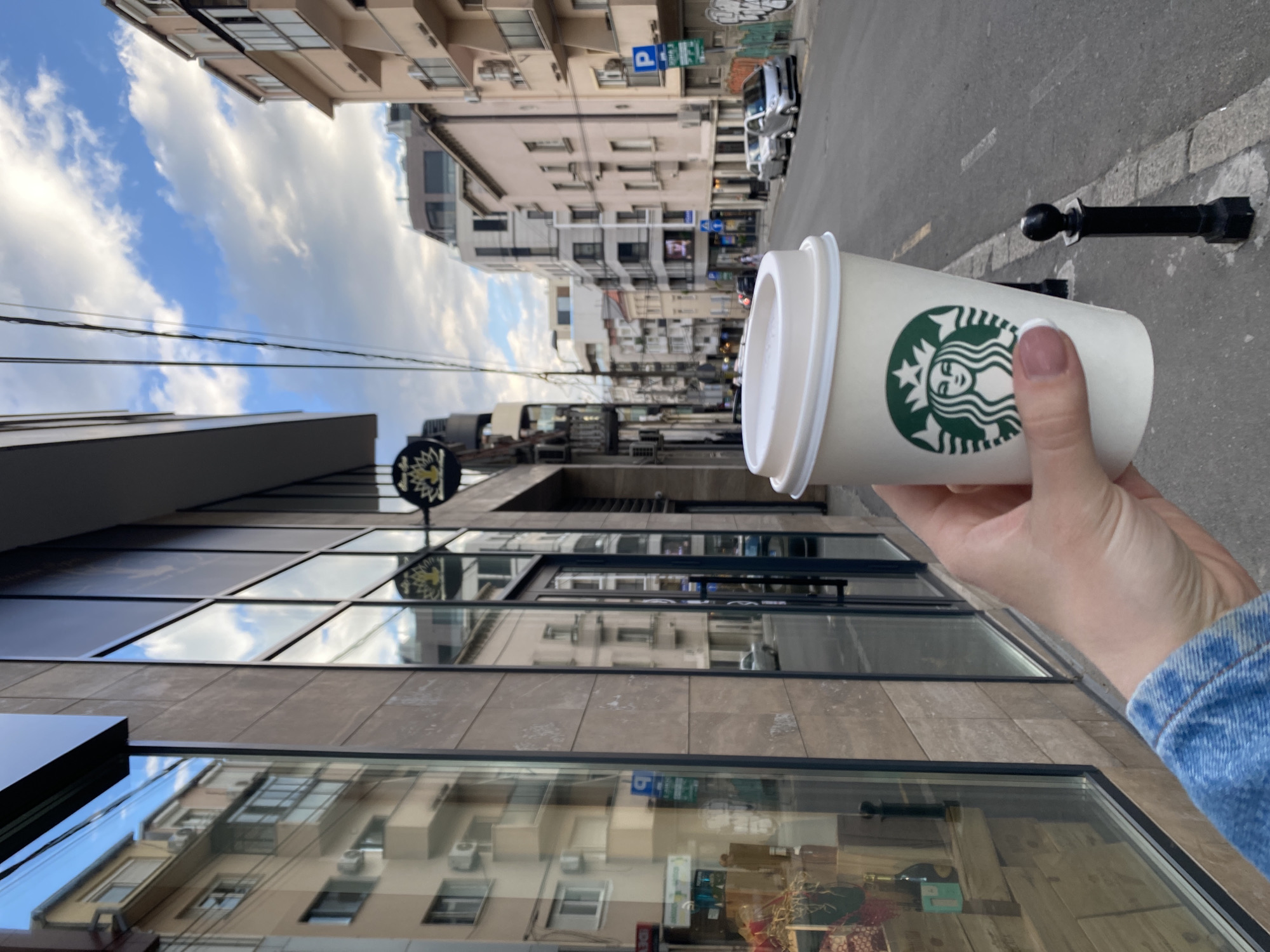 The image shows a person holding a Starbucks coffee cup in Belgrade, Serbia. The Starbucks logo is visible on the cup. The background includes buildings lining a street, with a clear sky and clouds overhead. A black bollard is seen on the edge of the sidewalk. The person is wearing a blue denim jacket. The buildings' reflections are visible in the glass windows of the Starbucks building. The street appears to be narrow with cars parked along the side. There is a parking sign in the distance.