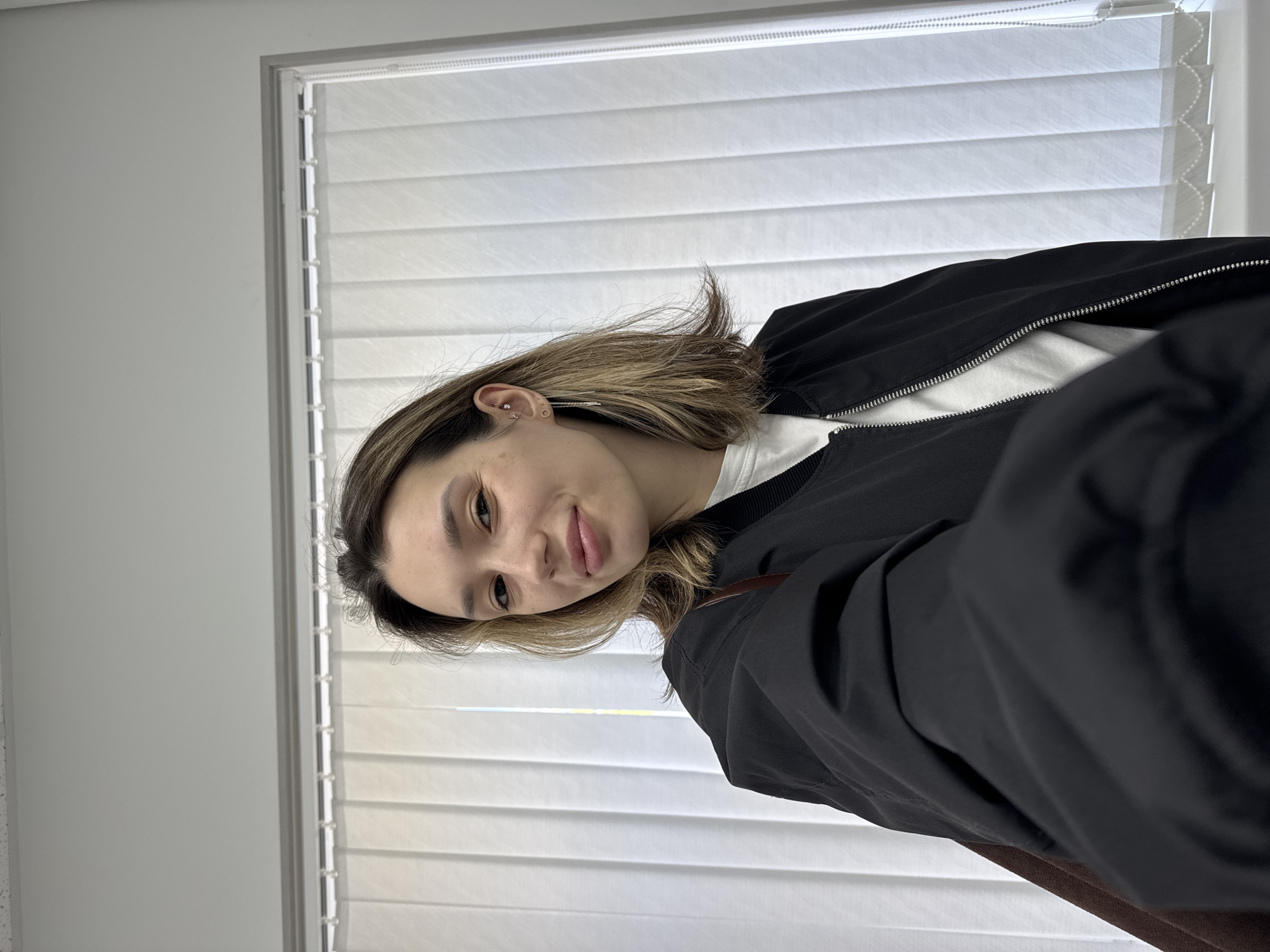 A young woman is captured in a close-up, indoor shot, looking directly at the camera with a soft smile. Her head and upper torso are visible, positioned against a backdrop of a window with white Venetian blinds and a light-colored wall. She has medium-length, straight hair, which is light brown with highlights, and some strands fall forward around her face. Freckles are visible on her cheek. On her visible ear, there are several piercings, including one on the helix and multiple on the lobe. She is wearing a black zippered bomber-style jacket over a white t-shirt, with the zipper partially open.
The background features a window with horizontal white blinds that allow diffused natural light into the room. The wall next to the window is a plain, light gray or white color. The lighting appears soft and even, highlighting her features. There is no discernible activity other than posing for the photo. No text is visible in the image. The overall mood is casual and pleasant.