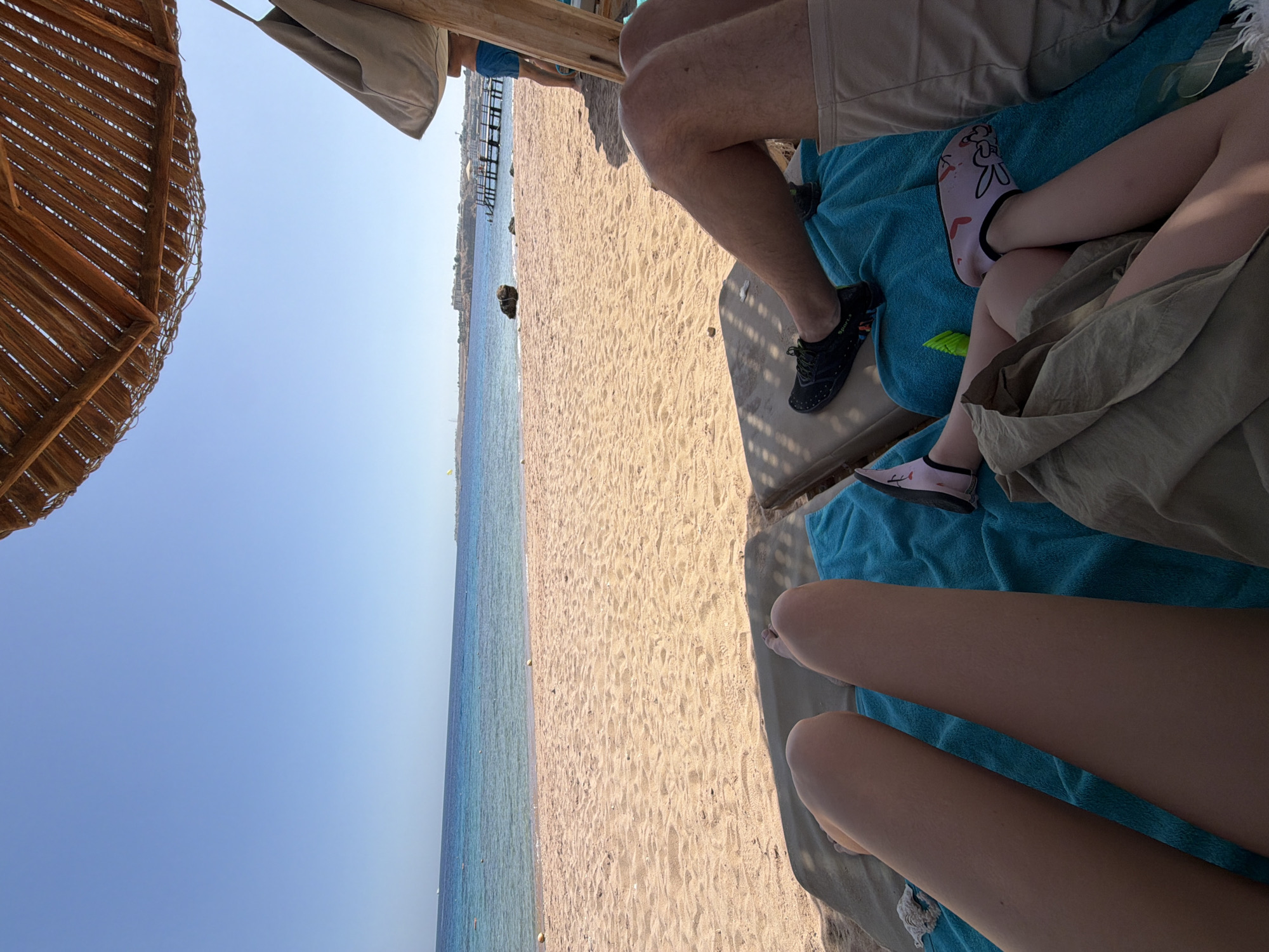 The image captures a vibrant beach scene, looking outwards from under the shade of a rustic, straw beach umbrella, partially visible in the top left. The perspective is from a seated position, showing the legs of at least three individuals relaxing on lounge chairs.
In the immediate foreground on the right, two pairs of legs are visible. The top pair, belonging to an adult, is tanned and clad in light-colored shorts and black water shoes that display "Sport.x" on the side. Below these, a second person's legs are seen, wearing light pink water socks adorned with a playful pattern, their lower body draped in a light-colored fabric, possibly a skirt or dress. A third pair of bare legs extends towards the bottom right corner, suggesting another individual is also relaxing. All are seated on lounge chairs covered with bright blue towels.
The midground features a wide, pristine sandy beach, gently sloping towards the clear, tranquil turquoise sea. The water appears calm, with light reflections, indicating a clear, sunny day. In the distance, along the coastline, a wooden pier or jetty extends into the water, and some structures or land are vaguely visible further along the shore. The sky is a brilliant, cloudless blue, meeting the horizon line where the sea stretches outwards.
The scene depicts a leisurely moment at a beach, likely a resort. The activity is one of relaxation and enjoying the seaside ambiance. The time of day appears to be sunny daytime, with bright light but some shade provided by the umbrella. The weather is excellent for a beach outing. This setting aligns with the provided location context of Sharm El Sheikh, Egypt, known for its beautiful beaches and resort environment.