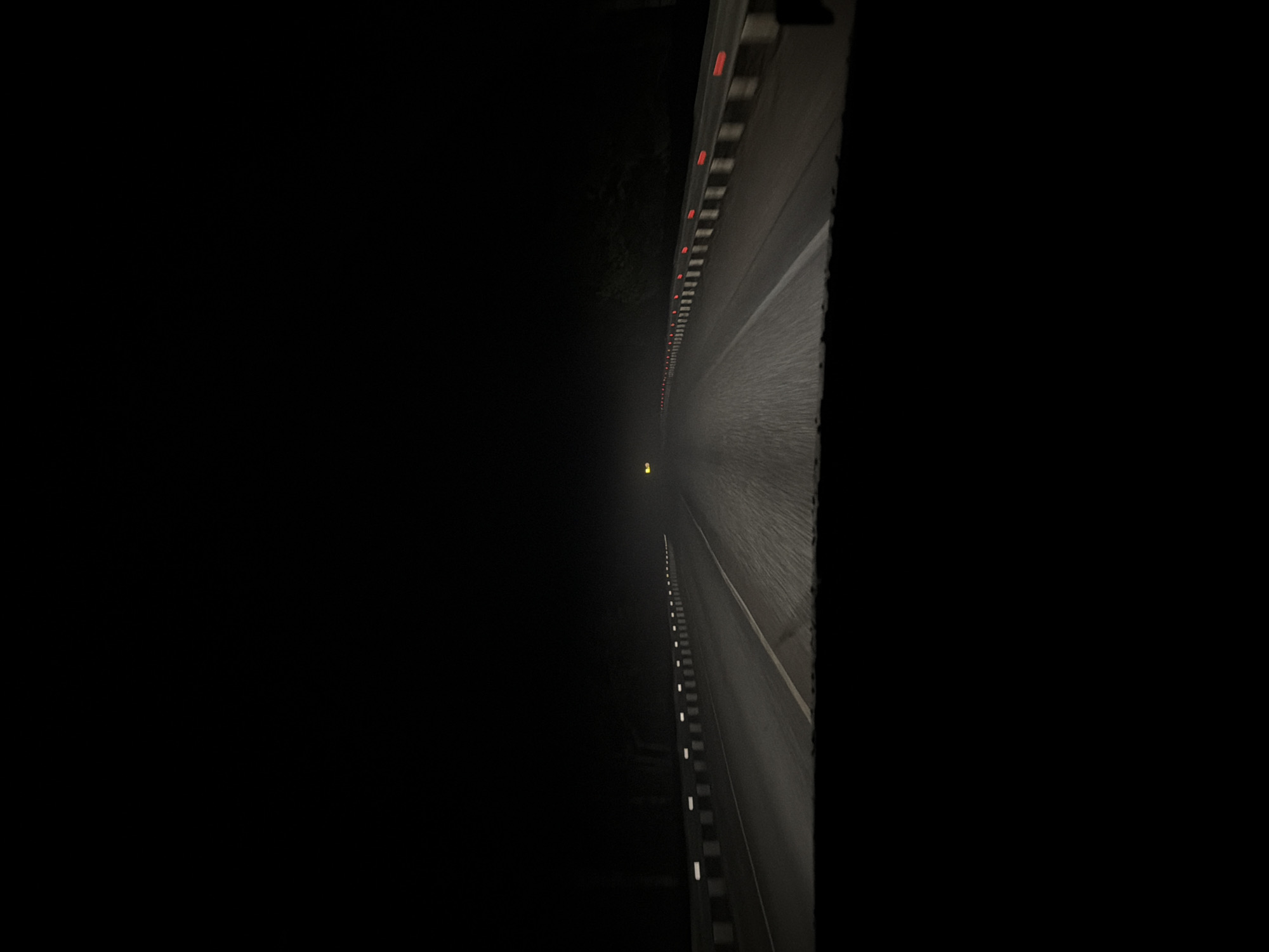 The image depicts a dark highway at night, possibly in fog. A single yellow light is visible in the distance, indicating a possible vehicle or source of illumination. Guardrails with alternating red and white reflectors line the sides of the road. The location is unknown.
