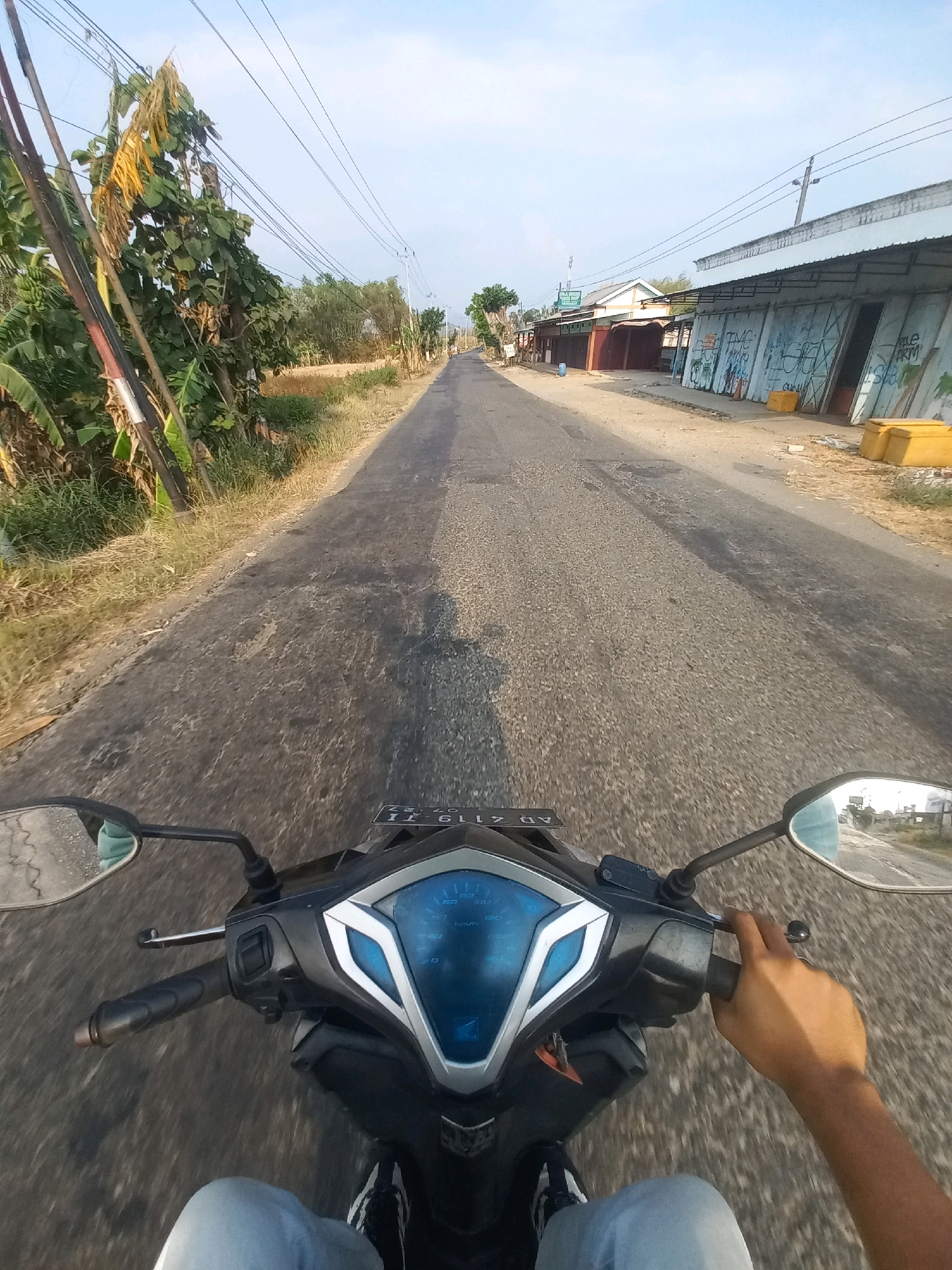 This is a first-person perspective, point-of-view shot from a person riding a motorcycle on a paved road in Baturetno, Indonesia. The road is in moderate condition with visible cracks and patches. On the left side of the road, there is dry grass, some leafy trees, and utility poles with numerous wires. On the right side of the road, there are several small, single-story buildings with tin roofs. Some of the buildings have graffiti on their corrugated metal shutters, and there are yellow objects, possibly containers, placed outside. The sky is clear with a light blue hue. The motorcycle's handlebars, dashboard with a blue digital display, a side mirror, and the rider's hand and legs are visible in the foreground. The rider's hand is on the handlebar, and their legs are clad in light blue jeans and black and white sneakers. The image captures a typical rural or semi-urban street scene during daylight.