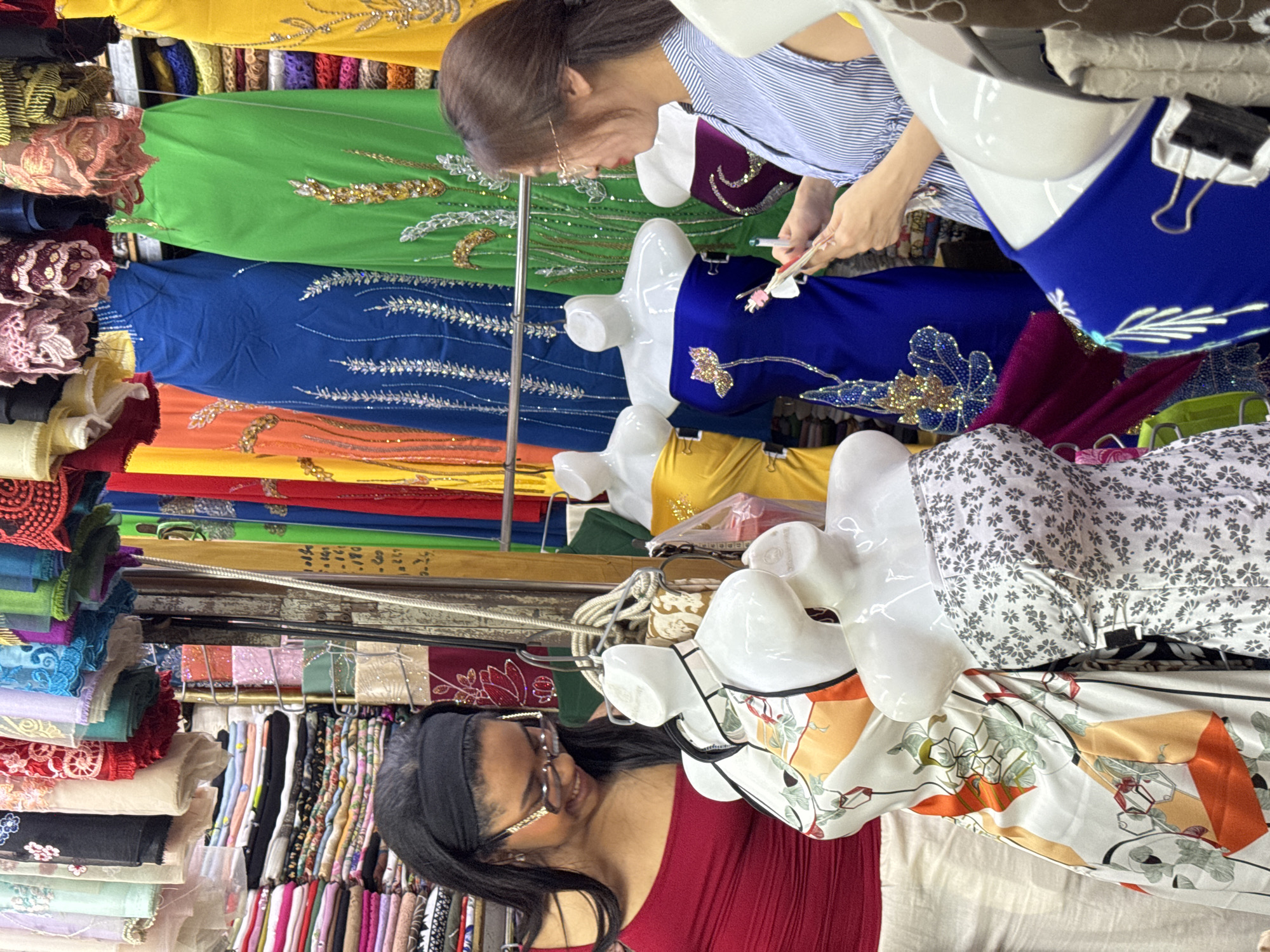The image captures a scene at Han Market in Đà Nẵng, Vietnam. A woman with short brown hair and glasses, wearing a blue and white striped shirt, is shown closely inspecting and possibly selecting fabrics. She appears to be holding a small object, perhaps a pen or tool, and a fabric swatch. Next to her are several mannequins displaying colorful, embroidered dresses. In the foreground, another woman with dark hair and glasses, wearing a red shirt, looks towards the dresses with a smile. Behind the women is a vibrant display of rolled textiles and fabrics hanging on racks, showcasing a wide variety of colors, patterns, and textures. The market appears crowded and bustling, reflecting a typical shopping environment.