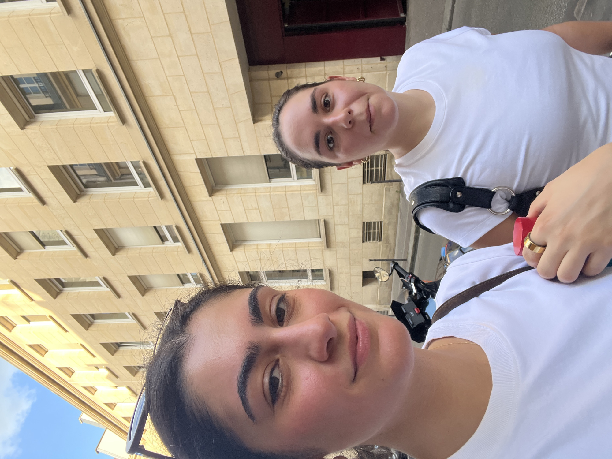 Two women are in the image. Both are wearing white t-shirts. The woman in the foreground has dark hair pulled back and is wearing sunglasses on her head. The woman in the background also has dark hair pulled back. She is wearing earrings. A black bag strap is visible on her shoulder. The background shows a beige building with windows, consistent with Parisian architecture. A motorcycle is also visible. The location is Paris, France.