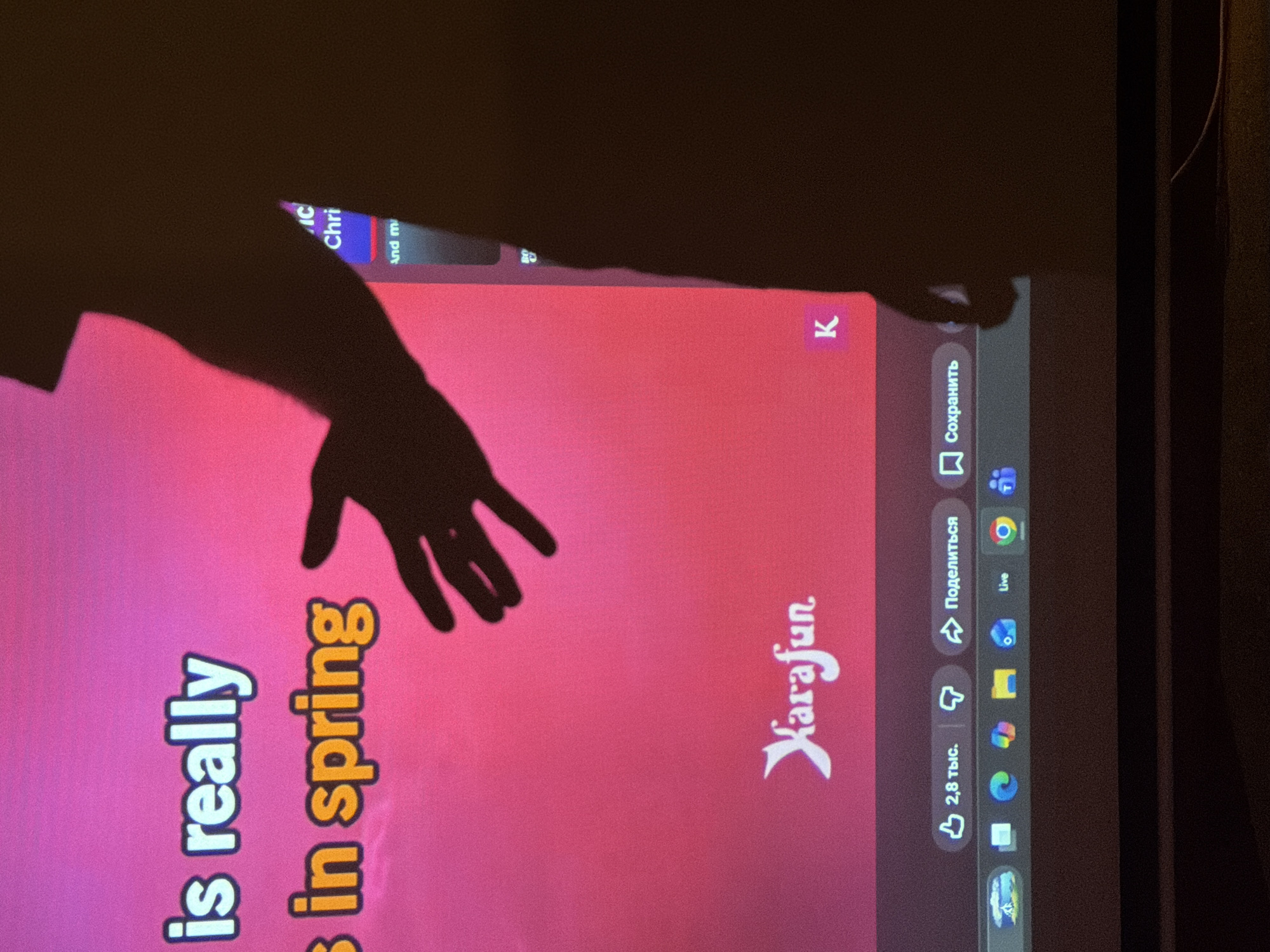 A close-up, indoor shot depicts a flat screen, possibly a monitor or television. The screen displays text in a gradient of pink: "is really" vertically aligned on the left, and "in spring" vertically on the right. Above these phrases is the word "Xarafun" in stylized script. Shadowy figures of a human arm and hand obscure the upper portion of the screen, suggesting someone is reaching towards it. On the right edge of the screen, a column of icons is visible with Russian text reading "Поделиться" and "Сохранить." Another text "В 2,8 тыс." is visible. At the top of the screen, "IC Chri" and "And m" is visible. The context suggests the screen may be displaying karaoke lyrics. The location in Yerevan, Armenia is not discernible from this image.