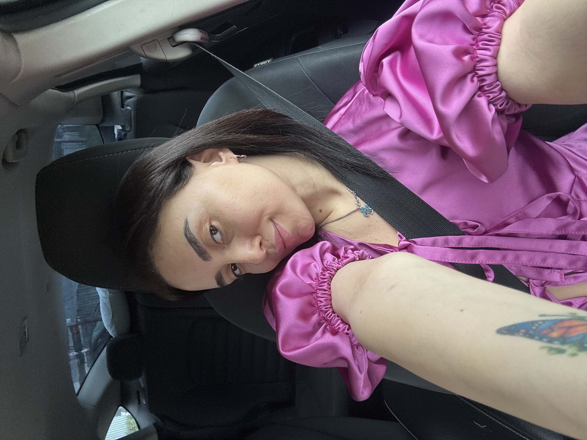 A woman is in the passenger seat of a car, wearing a purple satin dress. She is wearing a seatbelt. A butterfly tattoo is visible on her left forearm. The location appears to be Tbilisi, Georgia, as a building visible through the car window contains signage in the Georgian script.