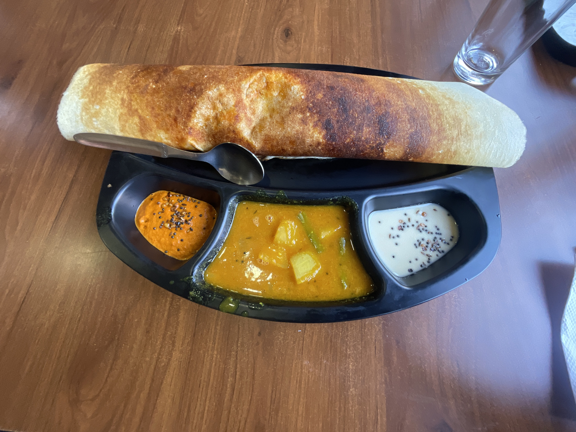 An overhead shot shows a close-up of a traditional South Indian meal, likely a dosa, served on a black segmented plate on a wooden table. The dosa, golden brown and rolled, lies to the right of the plate. The plate itself contains three distinct compartments filled with accompaniments. The top compartment holds a vibrant orange chutney with visible seeds. The middle compartment contains a yellow curry with potato chunks and green beans. The bottom compartment features a white chutney, also speckled with seeds. A silver spoon rests on top of the dosa, angled slightly. In the lower right corner, the rim of a clear glass is visible. The overall setting suggests a dining experience in Sidhpur, India, a city known for its rich cultural heritage and culinary traditions.