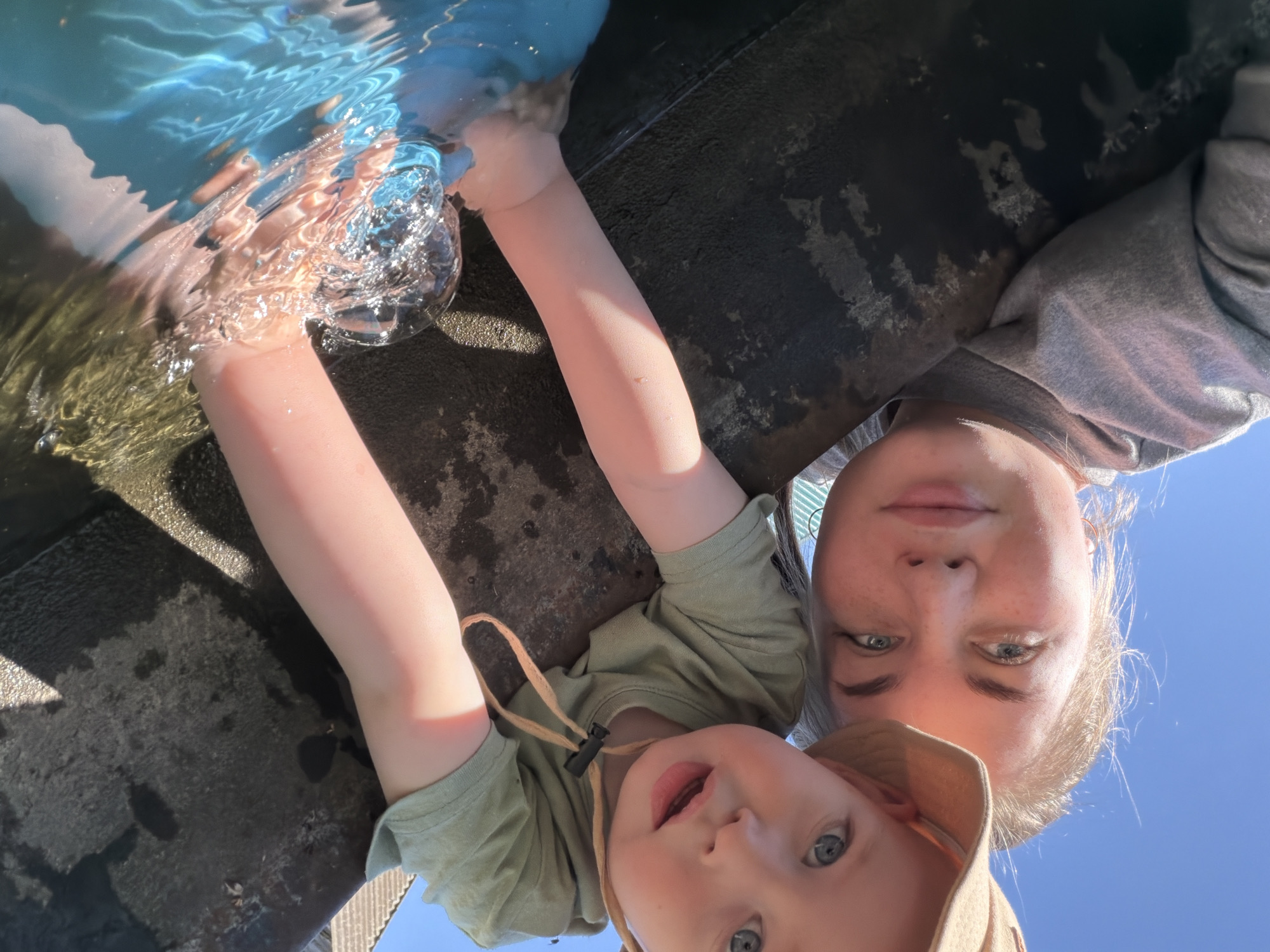 A tilted, close-up shot shows a woman and a young child, likely near or in water. The child, wearing a light green shirt and a tan hat with a strap, is looking upwards with their mouth slightly open. The woman, with blonde hair, is positioned behind and above the child, also looking upwards. One of the child’s arms is in clear water, with the hand submerged. The water's surface reflects light and sky. To the side, a dark, possibly concrete, surface borders the water. Clear blue sky is visible in the upper right of the frame. The exact location in Уральское, Russia is not ascertainable from the image alone.