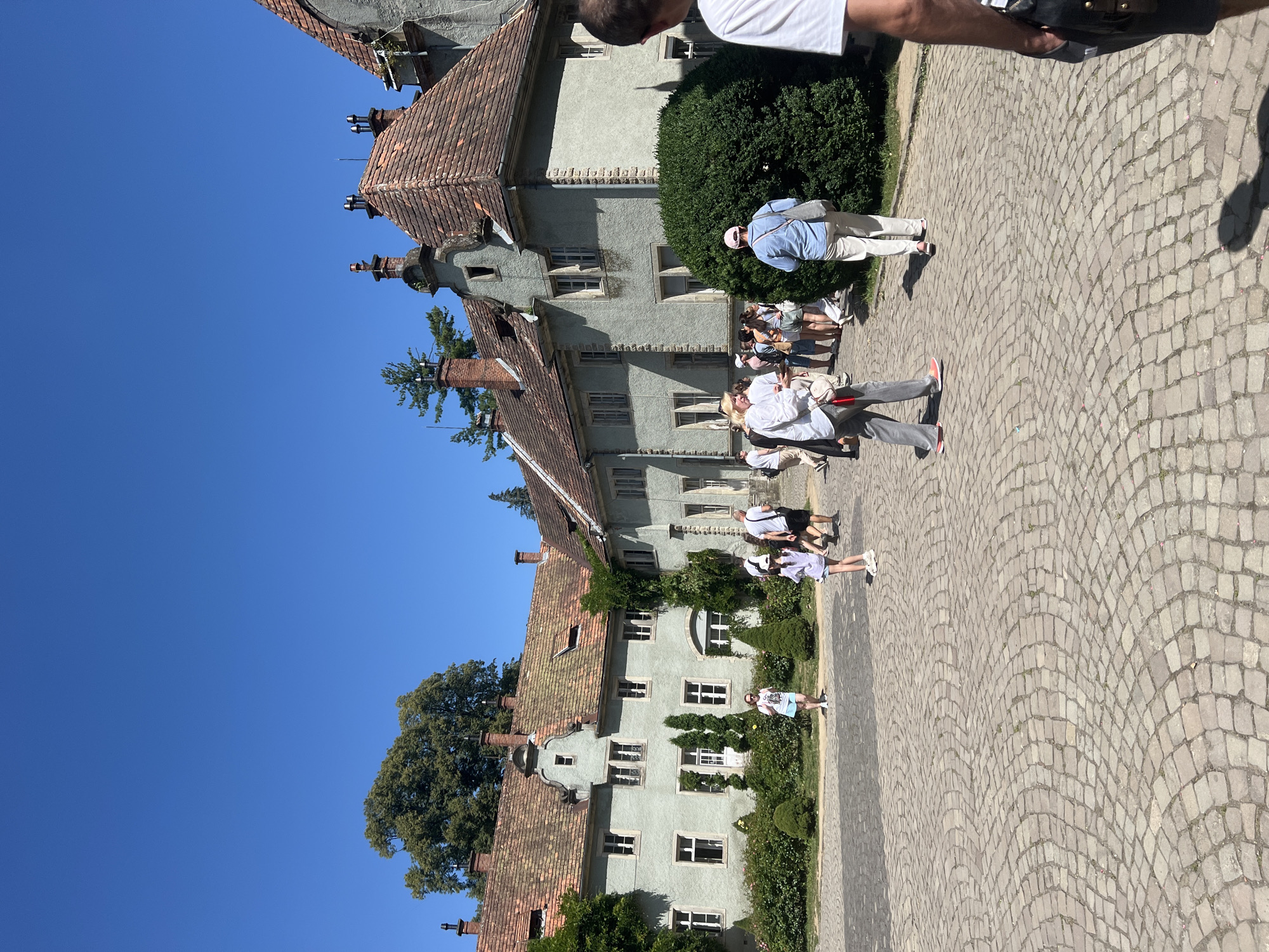 The image captures a sunny day in front of a historic castle in Karpaty, Ukraine. A group of tourists, dressed in casual summer attire, are seen walking across a cobblestone courtyard. The castle itself is a large, light green building with a terracotta tiled roof, featuring multiple chimneys and ornate architectural details. Lush green bushes and trees surround the building, adding to the picturesque scene. The sky is a clear, vibrant blue, indicating good weather. The overall atmosphere suggests a tourist destination, possibly a historical landmark open for public viewing.