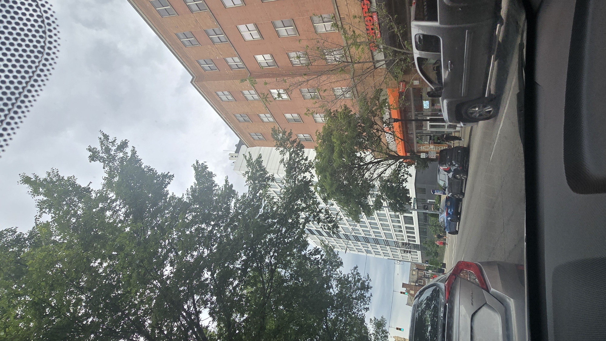 The image depicts a street scene in Cincinnati, Ohio, on a cloudy day. The perspective is from inside a vehicle, looking out through the windshield.
Dominating the left side of the frame are the lush green leaves of a large tree, partially obscuring the view of buildings. Behind the tree, a red brick building with numerous windows rises several stories. Adjacent to it, and further to the right, is a taller, modern white building with a grid-like pattern of windows.
The street below shows several parked cars and vehicles in motion, indicating typical urban traffic. A silver pickup truck is prominently visible in the middle ground, along with several other cars further down the street, including a blue sedan and darker SUVs.
Signs are visible on the buildings, but most are not clearly legible. One sign on the red brick building appears to have orange and black lettering. Another sign on a storefront below it has some text that is difficult to decipher. There are traffic lights visible, suggesting an intersection.
The weather appears overcast, as indicated by the grey sky and diffused light, suggesting it might be morning or afternoon. The overall mood is one of everyday urban life in a city. The scene is rich in architectural diversity, from older brick structures to contemporary designs, interspersed with the natural element of a large tree.