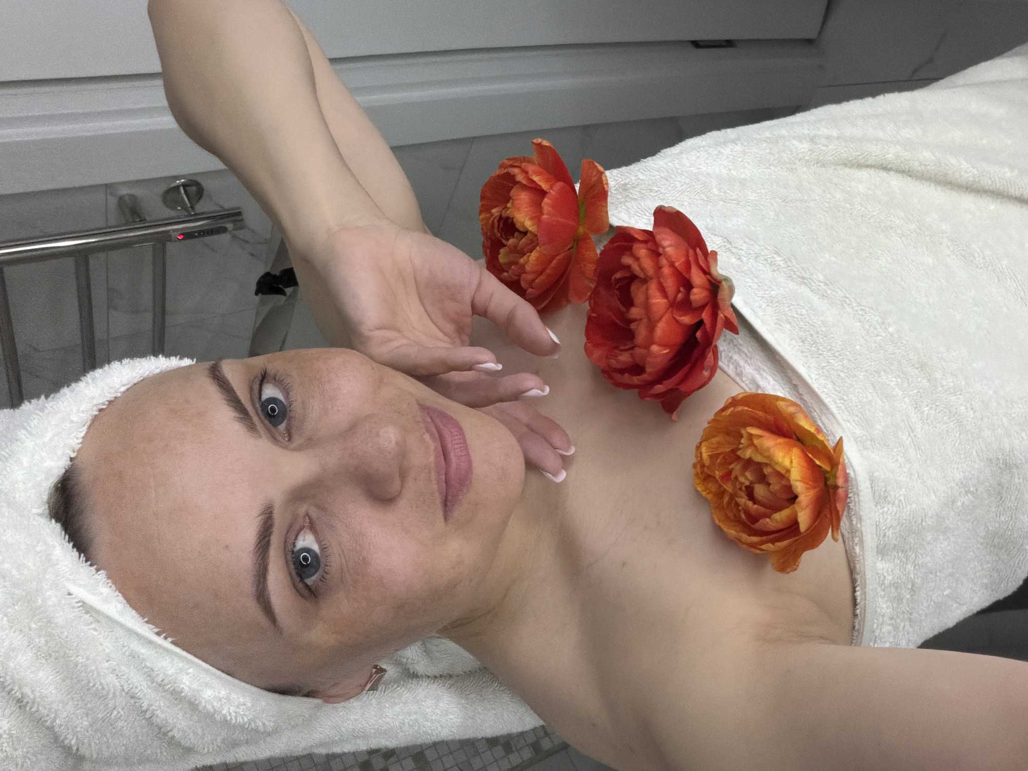 A woman lies on a white towel-covered surface, likely a massage or spa bed, with a white towel wrapped around her head. Three orange-red flowers are placed on her chest. Her left hand is raised near the flowers. The background includes what appears to be a white wall and a chrome towel rack. The location cannot be verified as Votkinsk, Russia with the information provided.