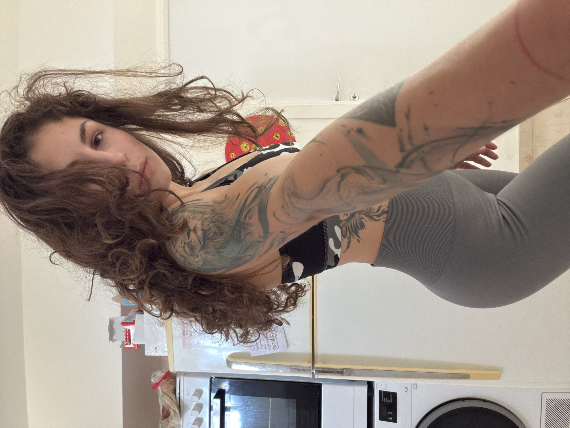 A young woman with long, curly brown hair and tattoos on her arm and torso is posing indoors in what appears to be a kitchen. She is wearing a sports bra and gray leggings. Behind her are kitchen appliances including an oven, refrigerator, and possibly a washing machine. The location is likely in Kiryat Bialik, Israel.