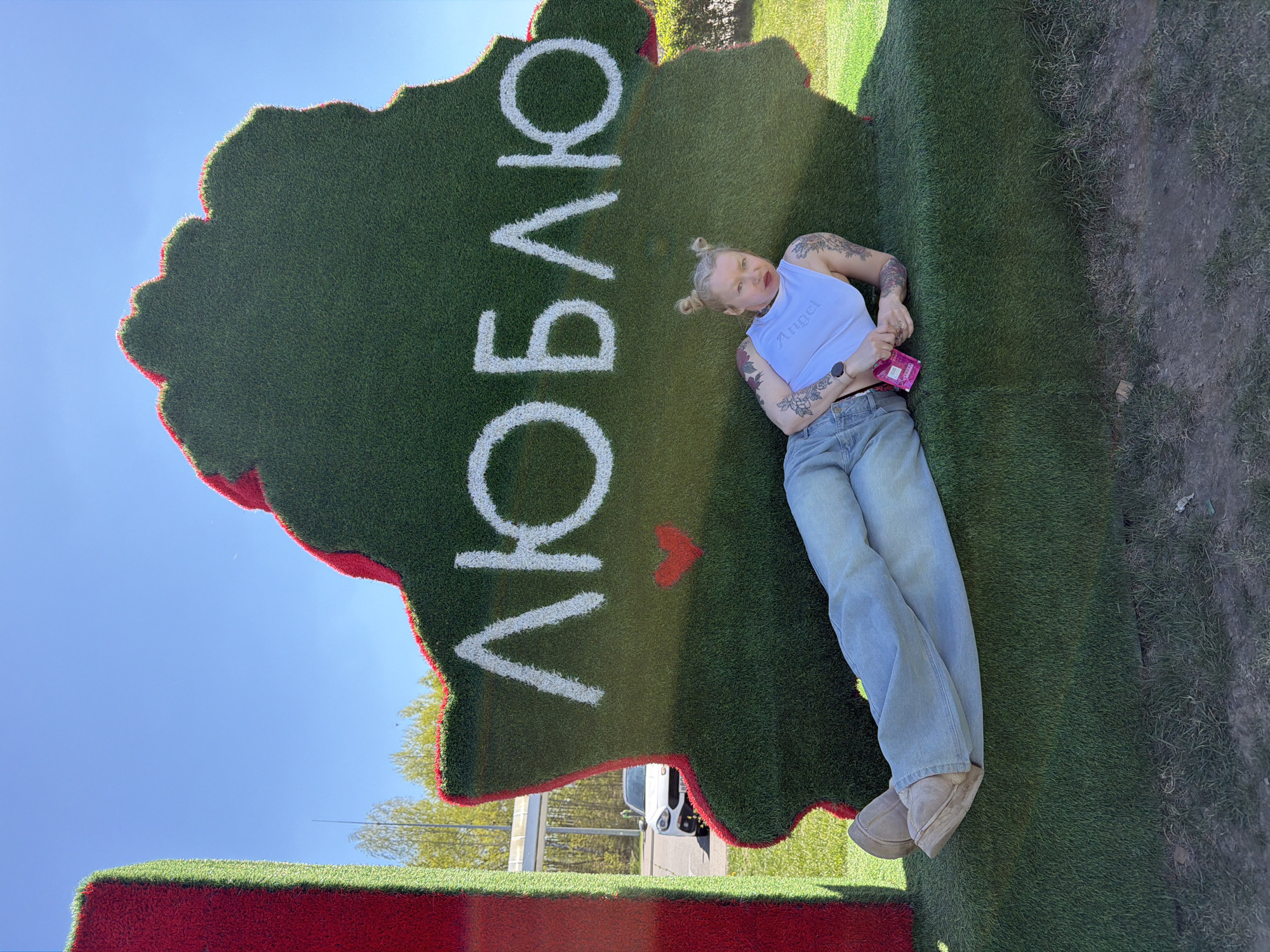 A woman with blonde hair styled in buns is lying on a large, green, map-shaped structure outlined in red, with the Cyrillic word "ЛЮБЛЮ" (meaning "I love" in Russian) spelled out in white. The structure, which appears to be covered in artificial grass, is sitting on a red base. The location is the city of Крошынскі сельскі Савет, Belarus. The woman is wearing a light blue top with the word "Angel" written on it, light blue jeans, and tan boots. She has tattoos on her arms. She is holding a pink object in her hands. The background shows a blue sky and some greenery. A white car is visible in the background.