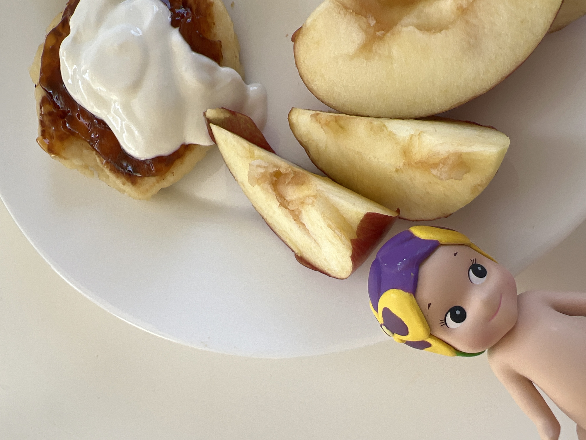 A white plate with food is displayed next to a Kewpie doll on a light-colored surface. The food consists of what appears to be a pancake with a layer of jam topped with white cream, and slices of a yellow-fleshed fruit, possibly a peach or nectarine. The Kewpie doll has light skin, black eyes, and hair that is painted purple and yellow. The surface is plain, without visible textures or details. There is no indication of the location in Budva, Montenegro.