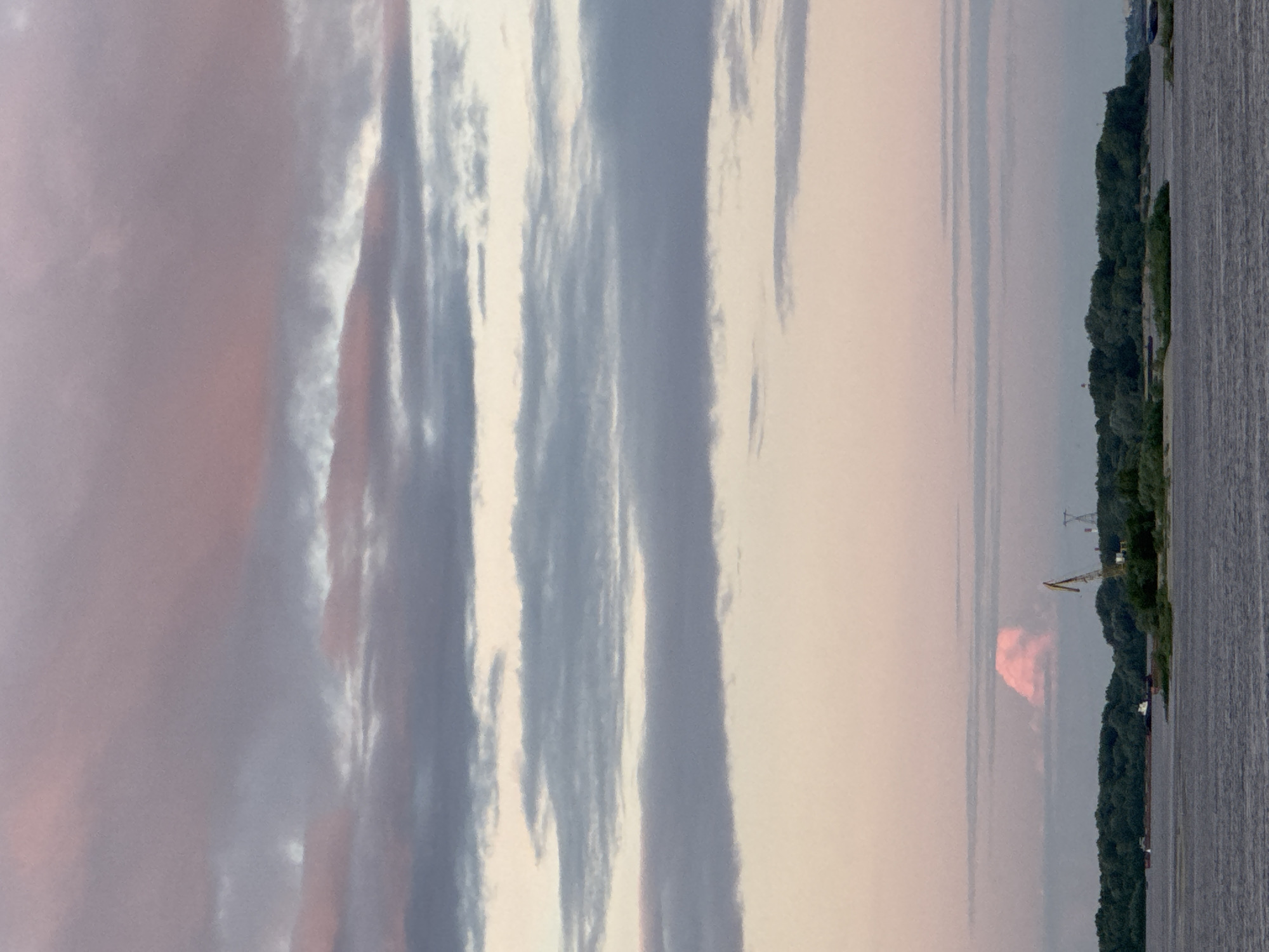 The image depicts a wide shot of a body of water, possibly a river, with a tree-lined shore on the right. The sky is filled with a mix of gray and pinkish clouds, suggesting either dawn or dusk. The water surface has a rippled texture, reflecting the muted colors of the sky.
On the shore, there is a dense line of trees, with a few structures visible amidst them. A crane, yellow in color, stands out against the greenery, hinting at industrial or construction activity. Power lines and poles are also visible further along the shoreline, indicating the presence of infrastructure. The overall impression is one of a serene, natural landscape with hints of human presence. The time of day appears to be either early morning or late evening, given the soft, diffused light and the colors in the sky. The weather seems calm, with no strong winds apparent on the water. The location context of Nizhny Novgorod, Russia, suggests this might be a view of the Volga River. No people are visible in the image, and there is no visible text.
