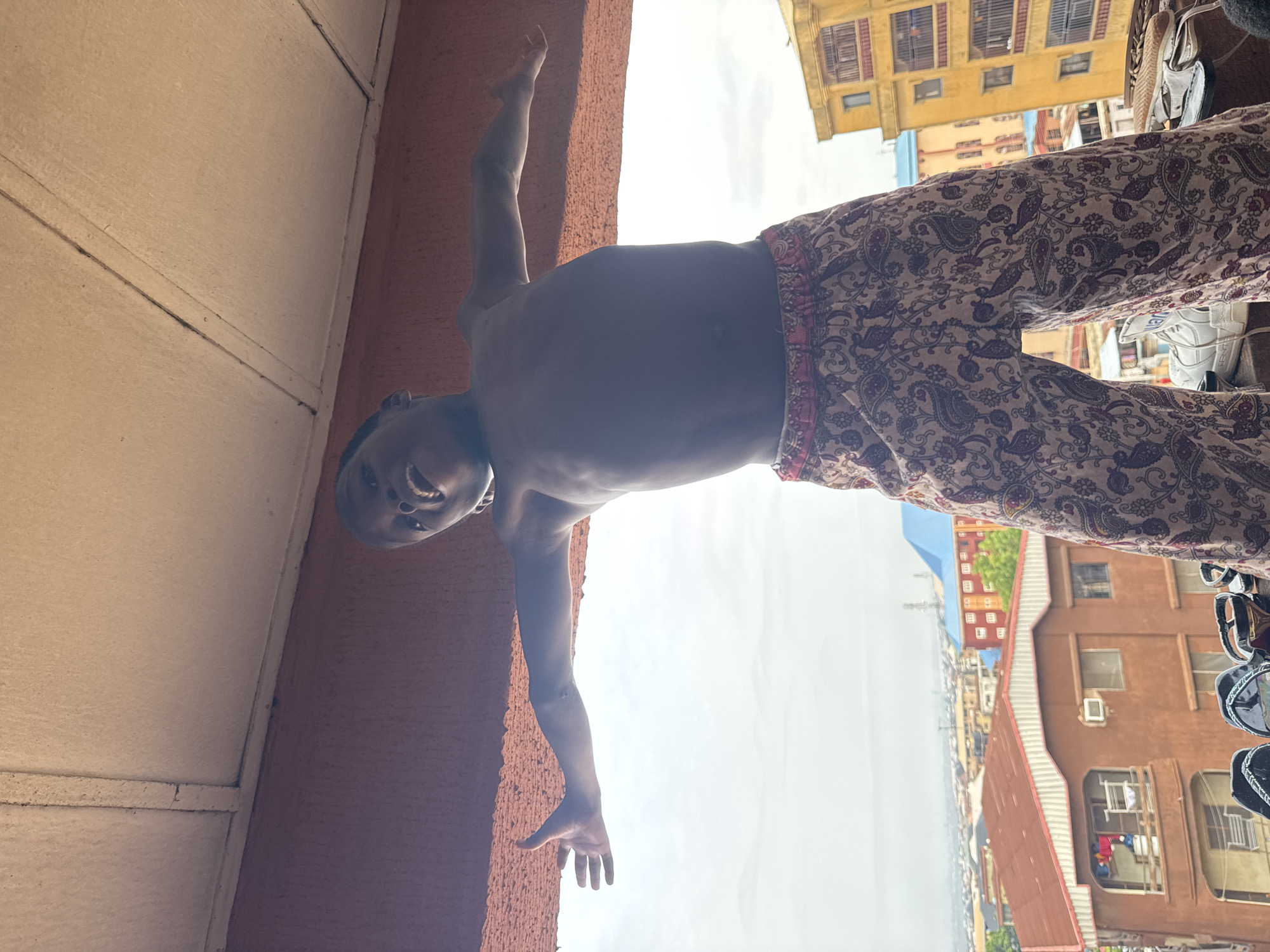 A low-angle medium shot of a shirtless young person wearing patterned pants, possibly pajamas, is positioned against a tan-painted wall. The subject is smiling, looking down towards the camera, with one arm extended upwards and the other hanging down. The background reveals a city landscape, identifiable as Onitsha, Nigeria, indicated by the architecture and urban environment. Multiple buildings are visible, and the sky is overcast.