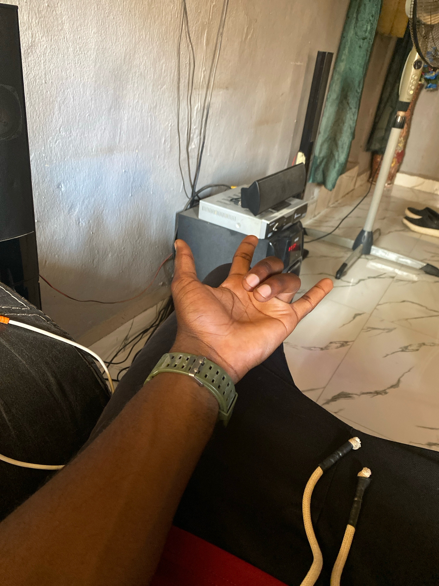 The image shows a person's left arm and hand, resting on black pants. The person is wearing a green watch on their left wrist. In the background, there is a white, textured wall with several black cables running down it. To the right of the cables, there is a grey electronic device with some smaller black devices stacked on top of it, one of which has a red light. Further to the right, a green cloth hangs, and a silver fan on a stand is visible. The floor is tiled with a marble pattern. The scene is indoors, likely in a residential setting in Jos, Nigeria, as indicated by the presence of common household electronics and the general interior design.