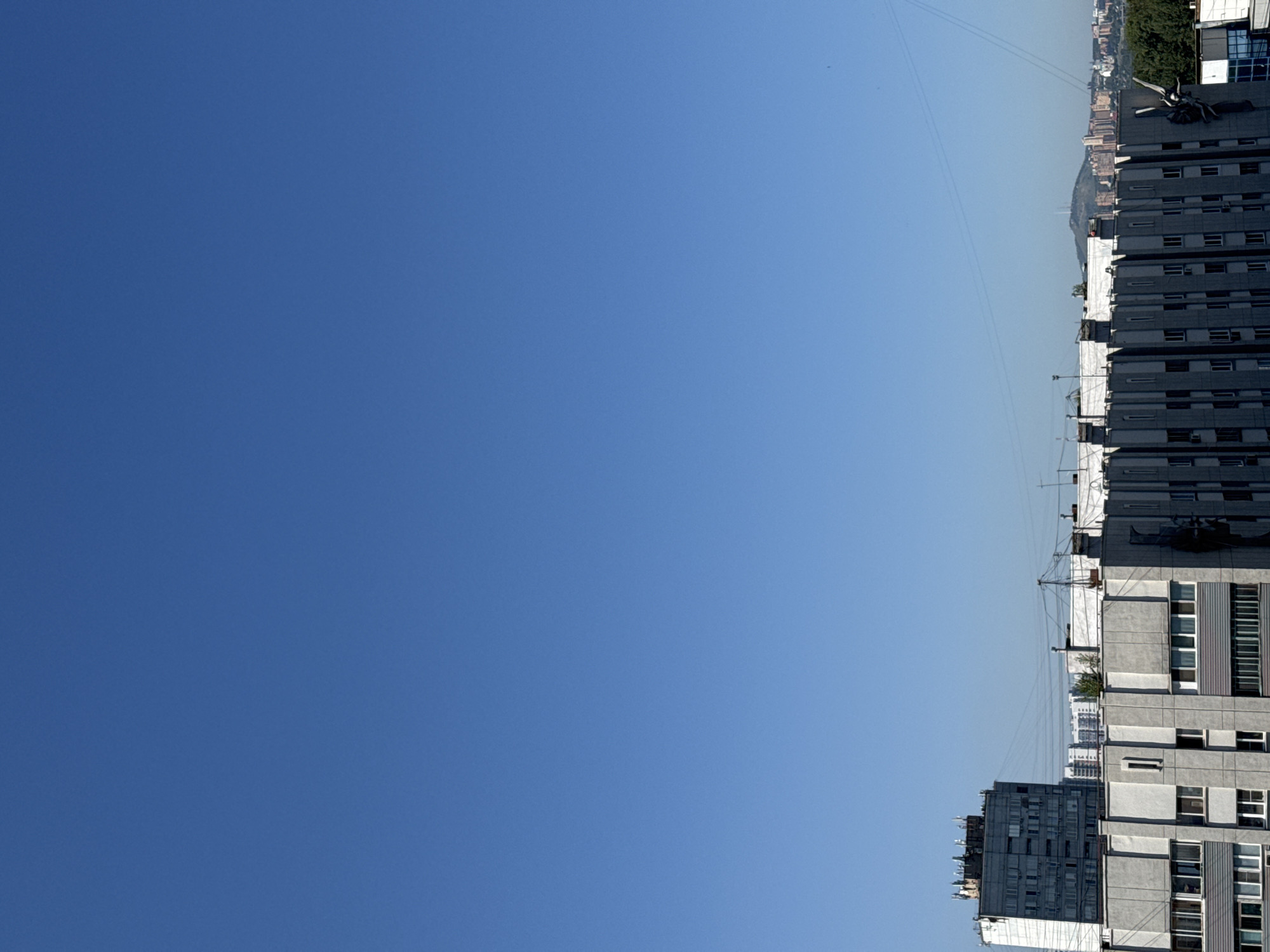 The image shows a view of urban buildings against a clear blue sky. The foreground features a large, grey, multi-story building with a geometric facade, characterized by vertical concrete panels and numerous windows. To the left, a taller, older, grey apartment building is visible, also with many windows and several antennas on its rooftop. In the middle ground, other buildings, some appearing residential, form a cityscape. The background reveals a distant, hazy landscape with hills and smaller buildings, suggesting a broader urban expanse. The scene is devoid of people or any visible activity, presenting a static snapshot of an urban environment. Considering the architectural style and the presence of distant hills, this scene is likely situated in Krasnoyarsk, Russia, a city known for its hilly terrain and Soviet-era apartment blocks.