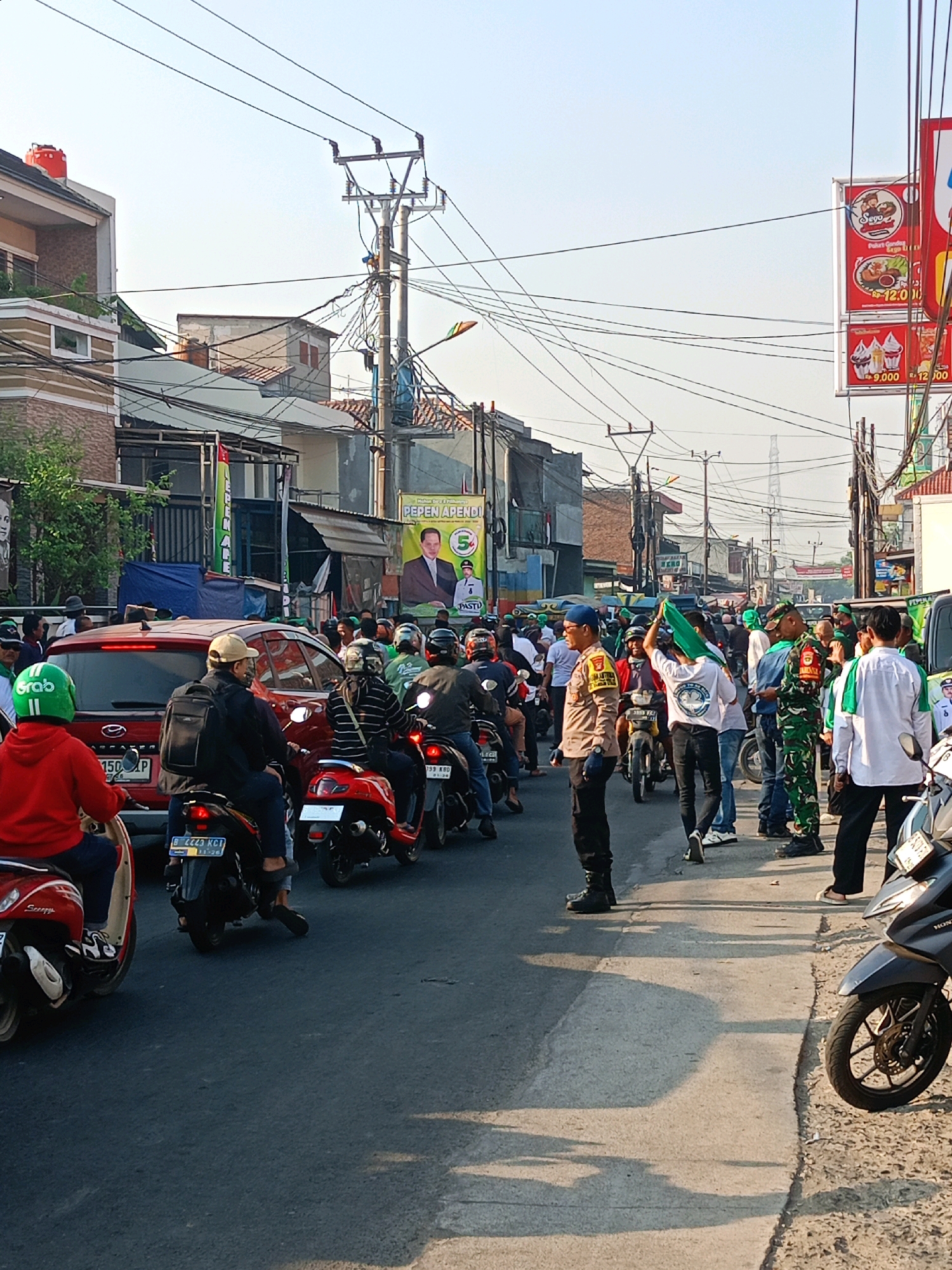The image depicts a busy street scene in Satriamekar, Indonesia, likely during daytime given the clear sky and shadows. A procession of motorbikes, cars, and pedestrians fills the road, suggesting a traffic jam or a planned event. Many individuals are wearing green clothing and some are holding green flags, possibly indicating a political rally or cultural celebration. A police officer in uniform is visible, standing in the middle of the road and observing the activity. The buildings lining the street appear to be a mix of residential and commercial properties, with some adorned with banners and advertisements. Utility poles with numerous electrical wires are prominent features of the urban landscape. The overall impression is one of a dynamic and somewhat congested urban environment.